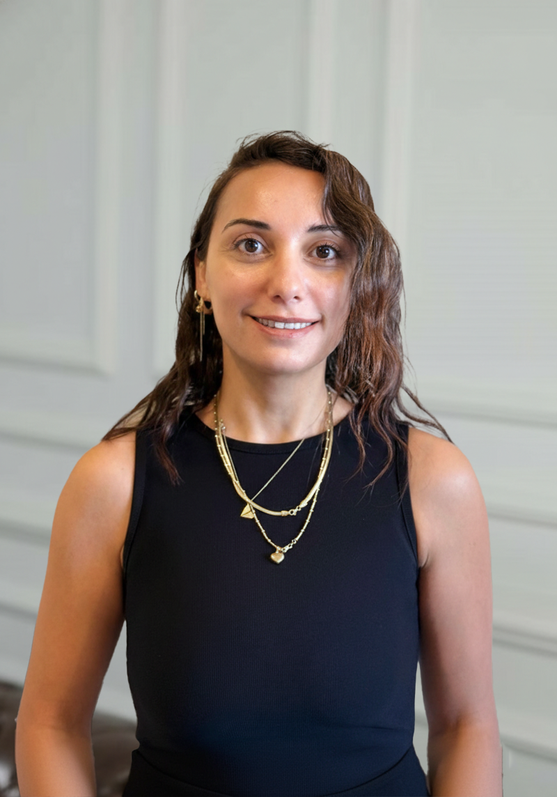 A woman with shoulder-length brown hair wearing a sleeveless black top, gold necklaces, and gold earrings, standing indoors against a light-colored wall.