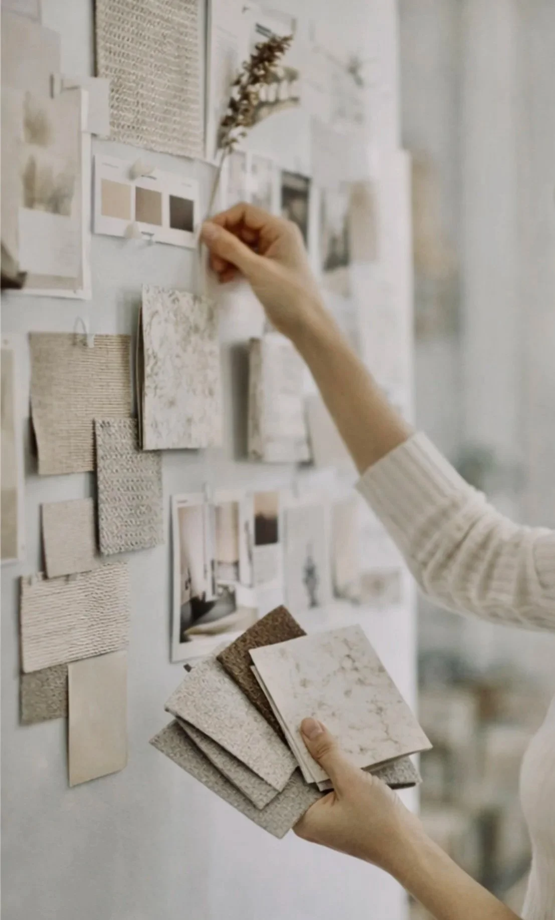 A person's hand holding a small stack of various textured sample tiles and another hand pinning a photo or sample to a wall decorated with other photos, samples, and swatches.