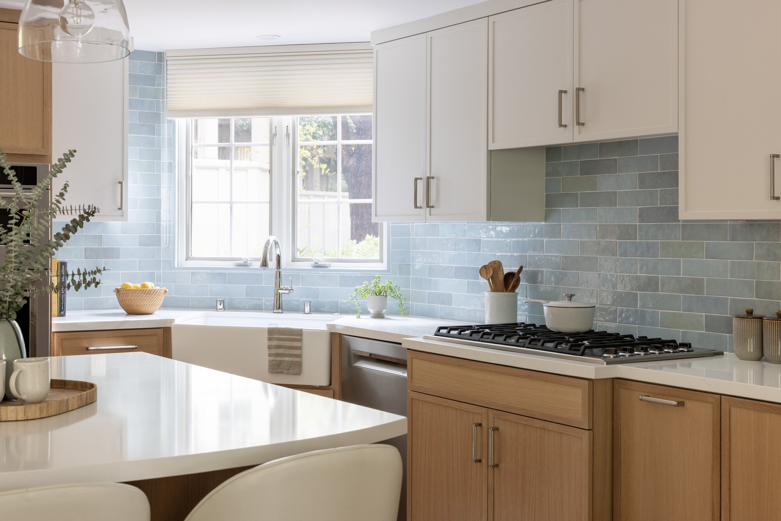 Kitchen with white cabinets, light blue tile backsplash, window above the sink, wooden lower cabinets, and a white countertop with cooking utensils and decor.