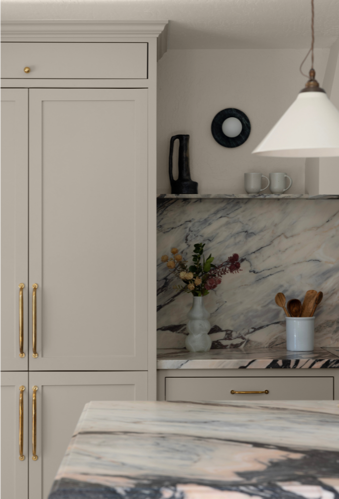 Kitchen with white cabinetry, gold handles, a marble backsplash, a marble countertop, a corner shelf with a black sculpture, two white mugs, a white vase with flowers, and wooden utensils in a white holder, with a hanging light fixture.