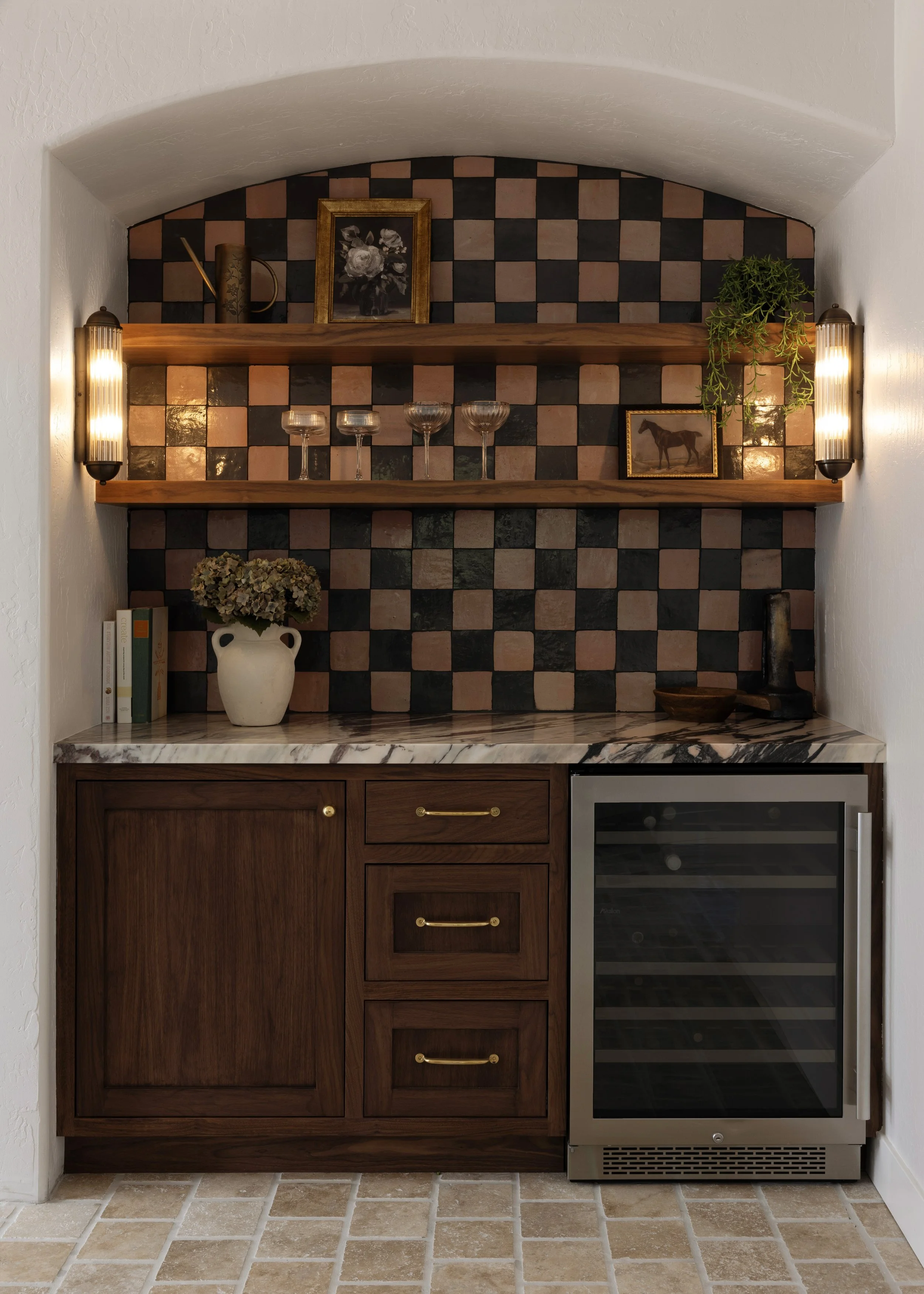 A small kitchen nook with a marble countertop, wooden cabinetry, a wine cooler, and a black and pink checkered tile backsplash. Floating wooden shelves hold glassware, books, a vase with dried flowers, and decorative items, with wall-mounted lights on each side of the nook.