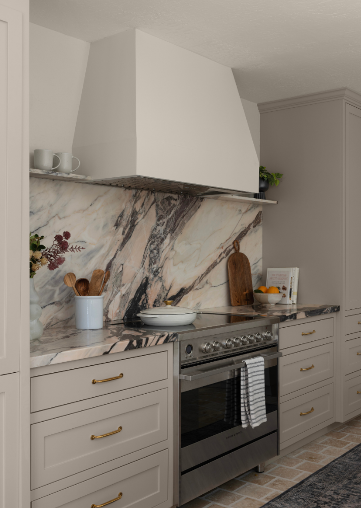 Kitchen with beige cabinetry, marble countertop, stainless steel oven, a marble backsplash, and decorative items including cutting boards, a fruit bowl, and a potted plant.