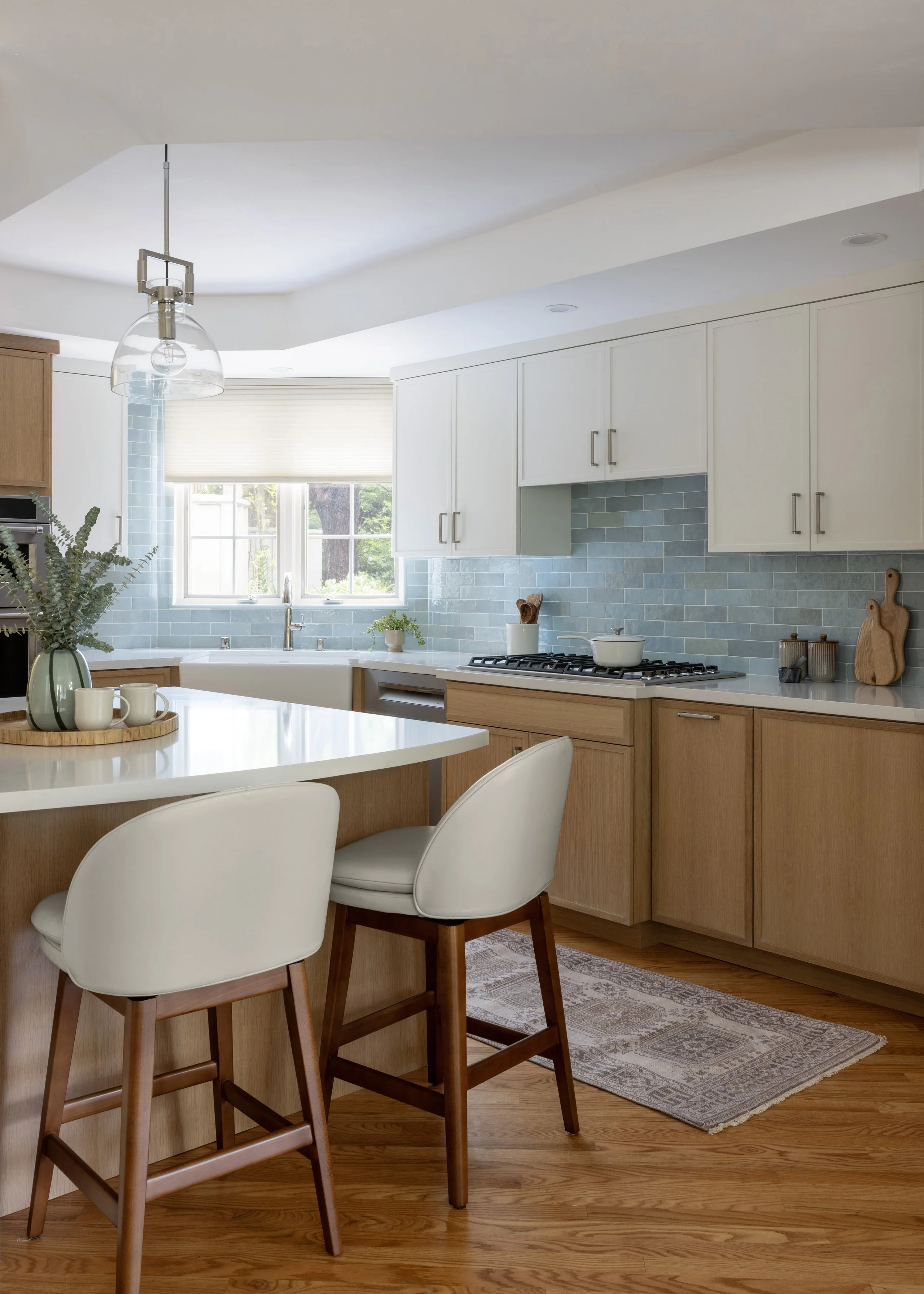 A modern kitchen with white cabinets, blue tile backsplash, wooden lower cabinets, and a white countertop. There is a window above the sink, natural light, and a kitchen island with seating, decorated with a vase of plants and mugs.