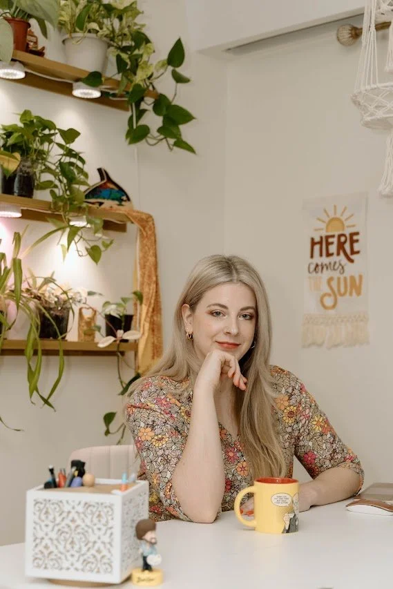 A woman with long blonde hair sitting at a white desk with a yellow mug, surrounded by plants and wall decorations.