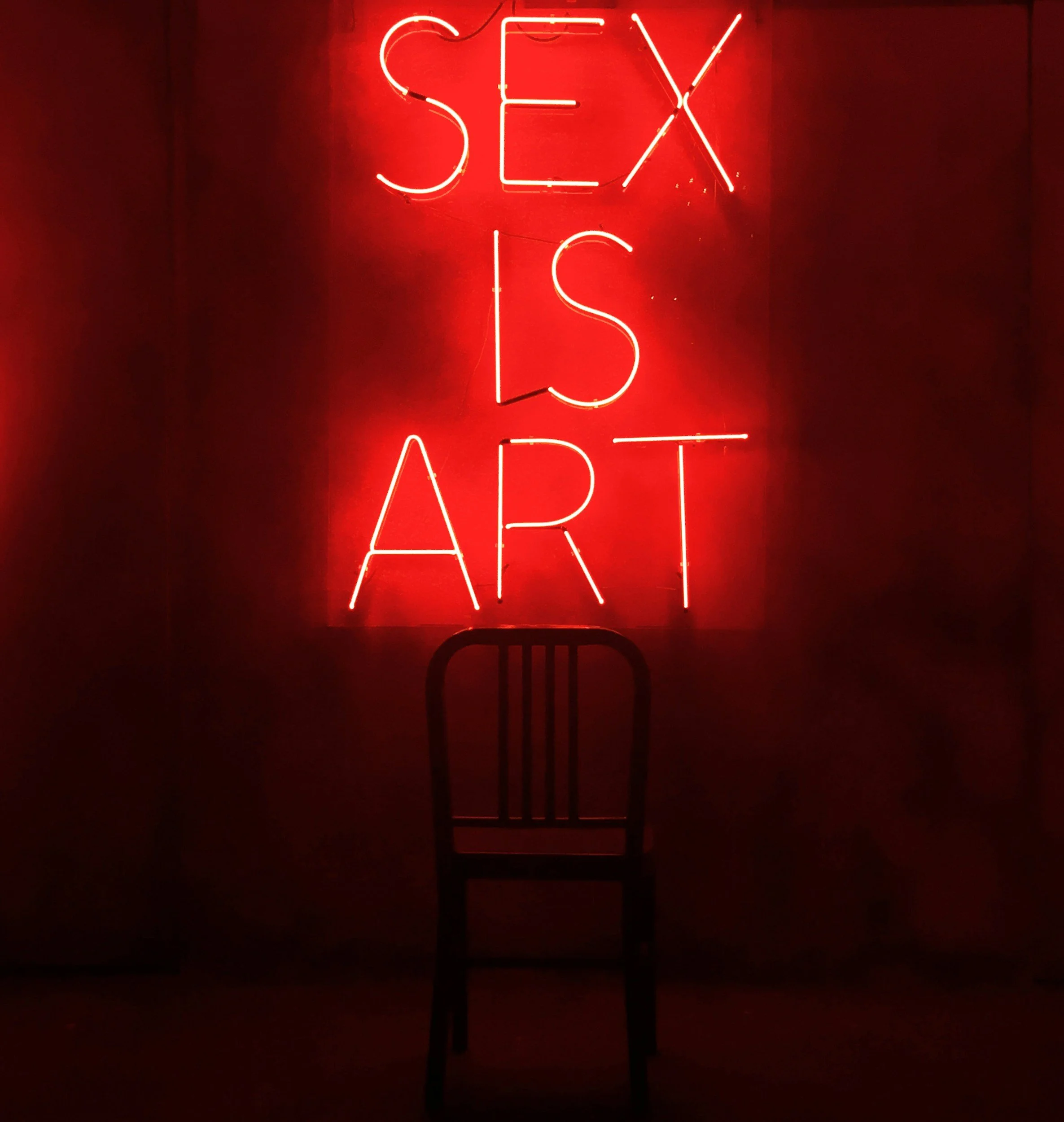 Red neon sign spelling out the words "SEX IS A ART" against a dark red wall.