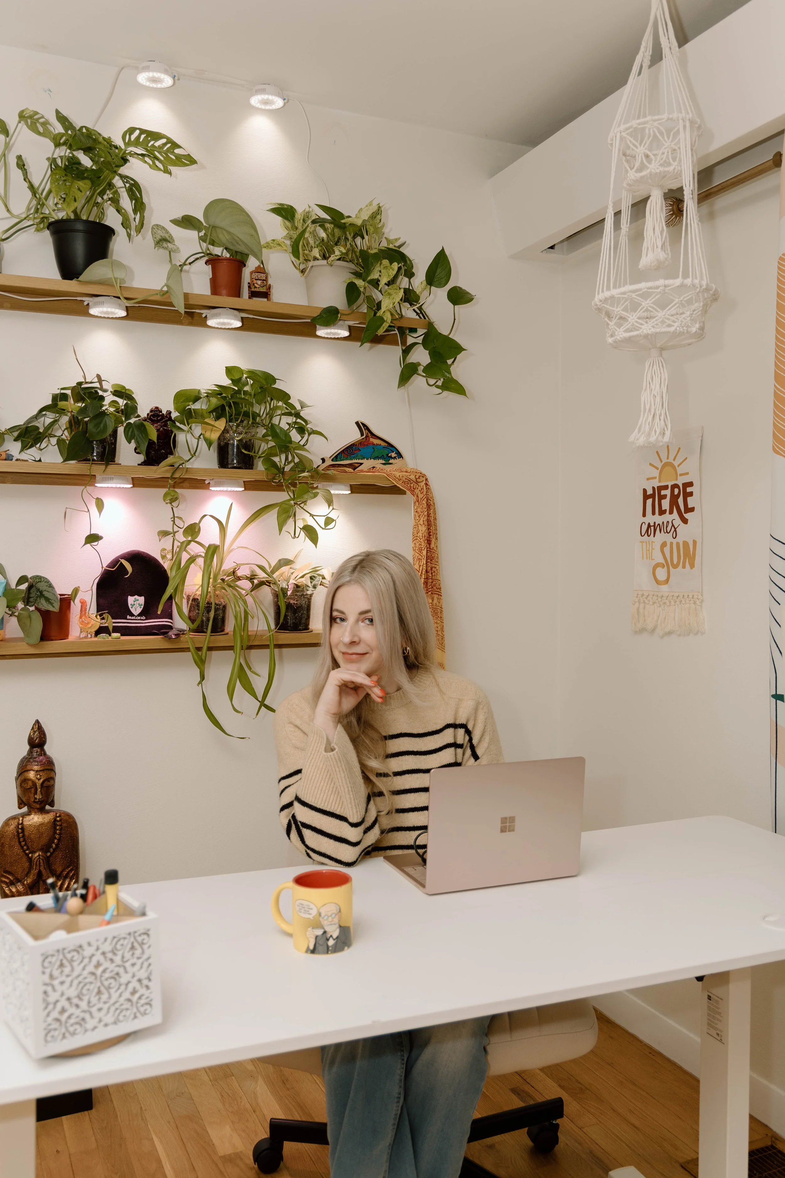 A woman sitting at a white desk with a laptop open in front of her, smiling and looking at the camera, surrounded by decorative items and houseplants in a cozy, well-lit room.