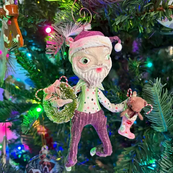 A Santa-themed Christmas tree ornament resembling a whimsical elf with a winking face, wearing a pink hat, polka-dot shirt, and striped pants, holding a small wreath in one hand and a tiny stocking with a teddy bear in the other.