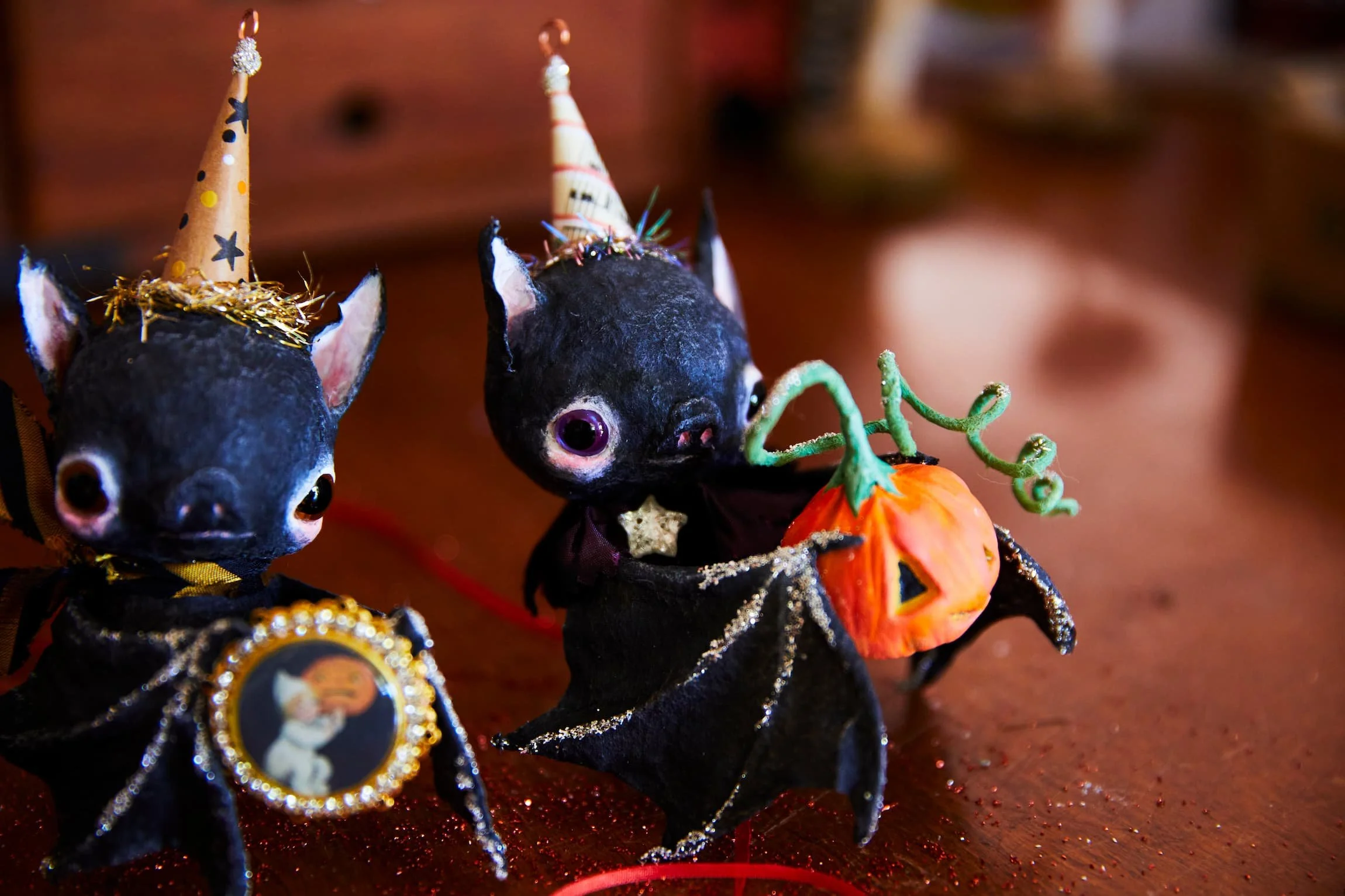 Decorative spun cotton miniature cats dressed in black outfits with gold trim and wearing party hats and medals, holding pumpkins, on a sparkly red surface.