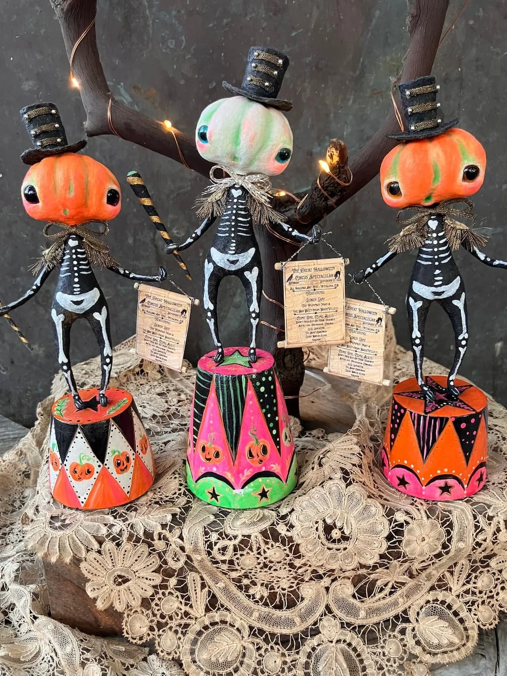 Three Halloween pumpkin and skeleton ornaments with painted faces, dressed in skeleton costumes, wearing top hats, and standing on decorated bases, displayed on a lace-covered surface.
