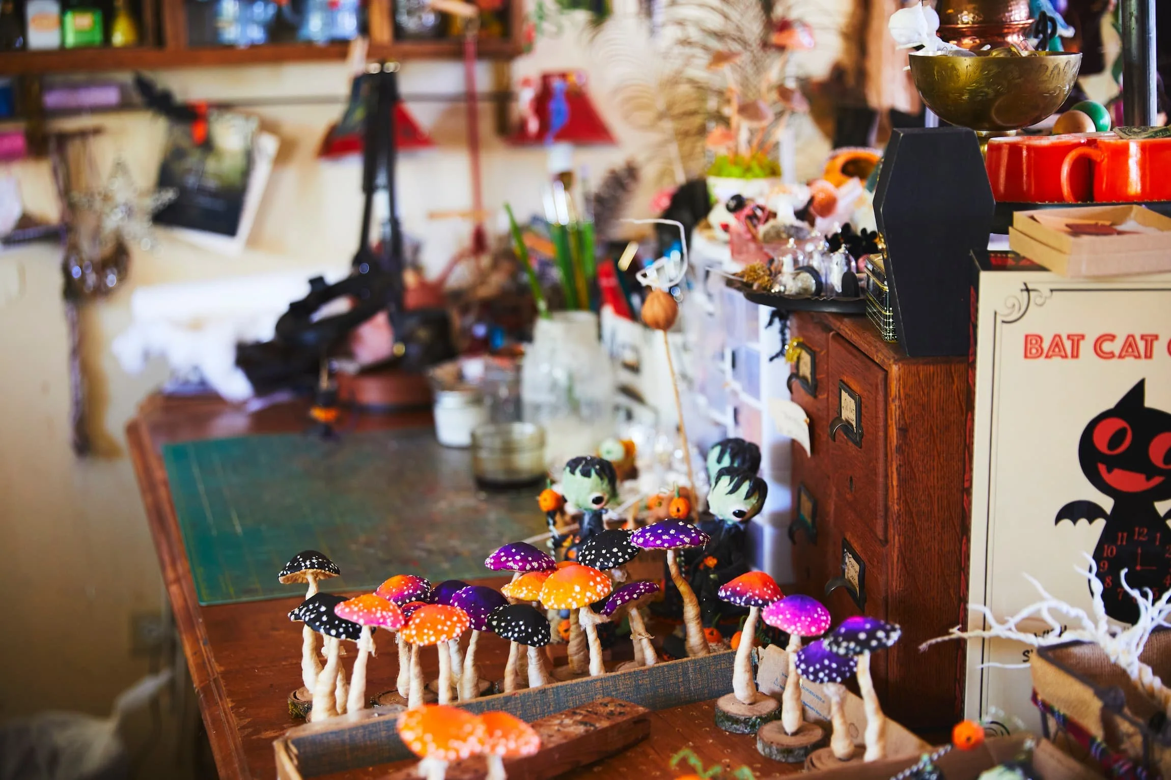Display of colorful handmade mushroom decorations arranged on a table, with various small figurines including characters with large eyes and orange accessories, inside a cozy store with shelves of miscellaneous items in the background.