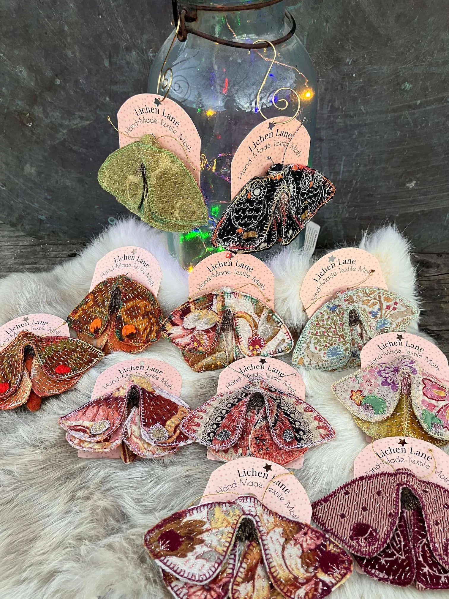 Display of embroidered textile butterflies attached to pink tags with 'Lichen Lane Hand-Made Textile Moth' printed on them, arranged on a fur surface with a glass jar in the background.