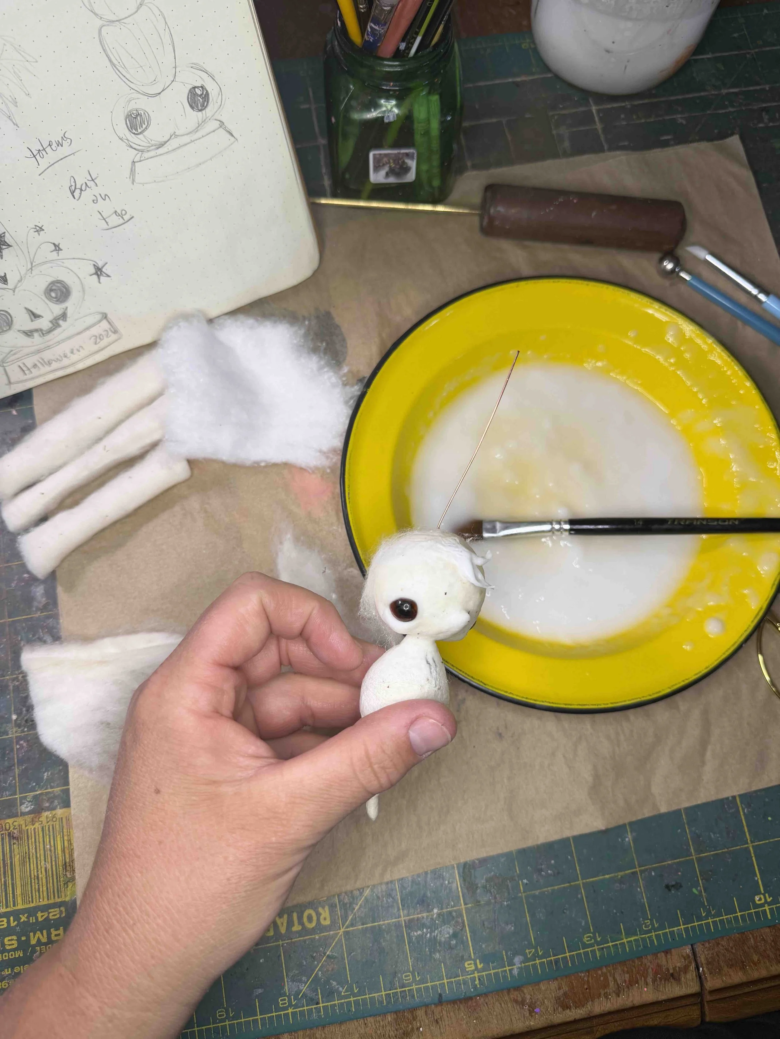 A person holding a small, white, fabric doll with large, black eyes. The background includes a yellow bowl with white liquid, a cutting mat with measurements, a jar with colored pencils, a roll of fabric, cotton stuffing, and a sketchbook with Hallow
