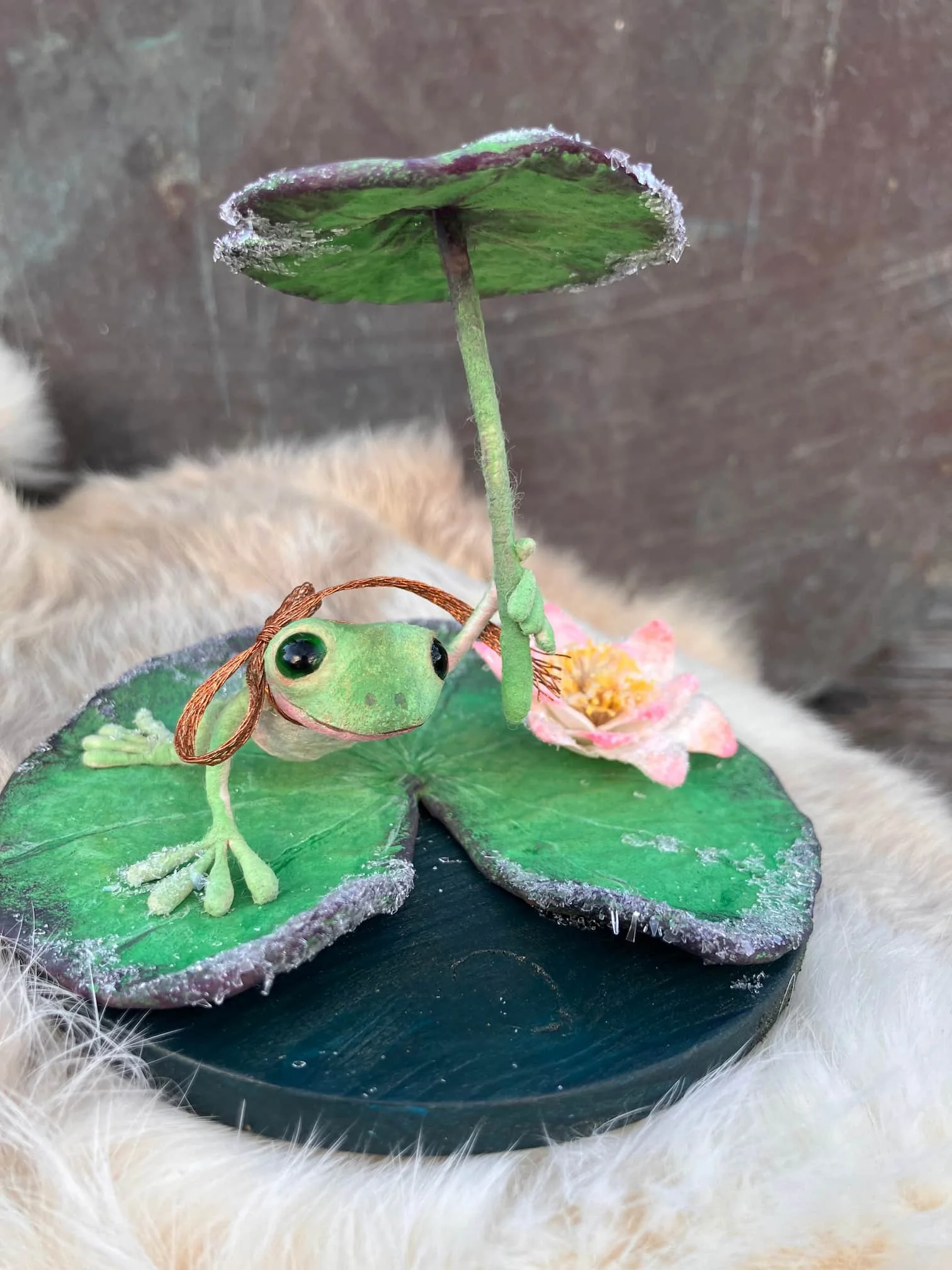 Decorative frog holding a lily pad and flower, placed on a round black base, surrounded by fluffy cream-colored material.