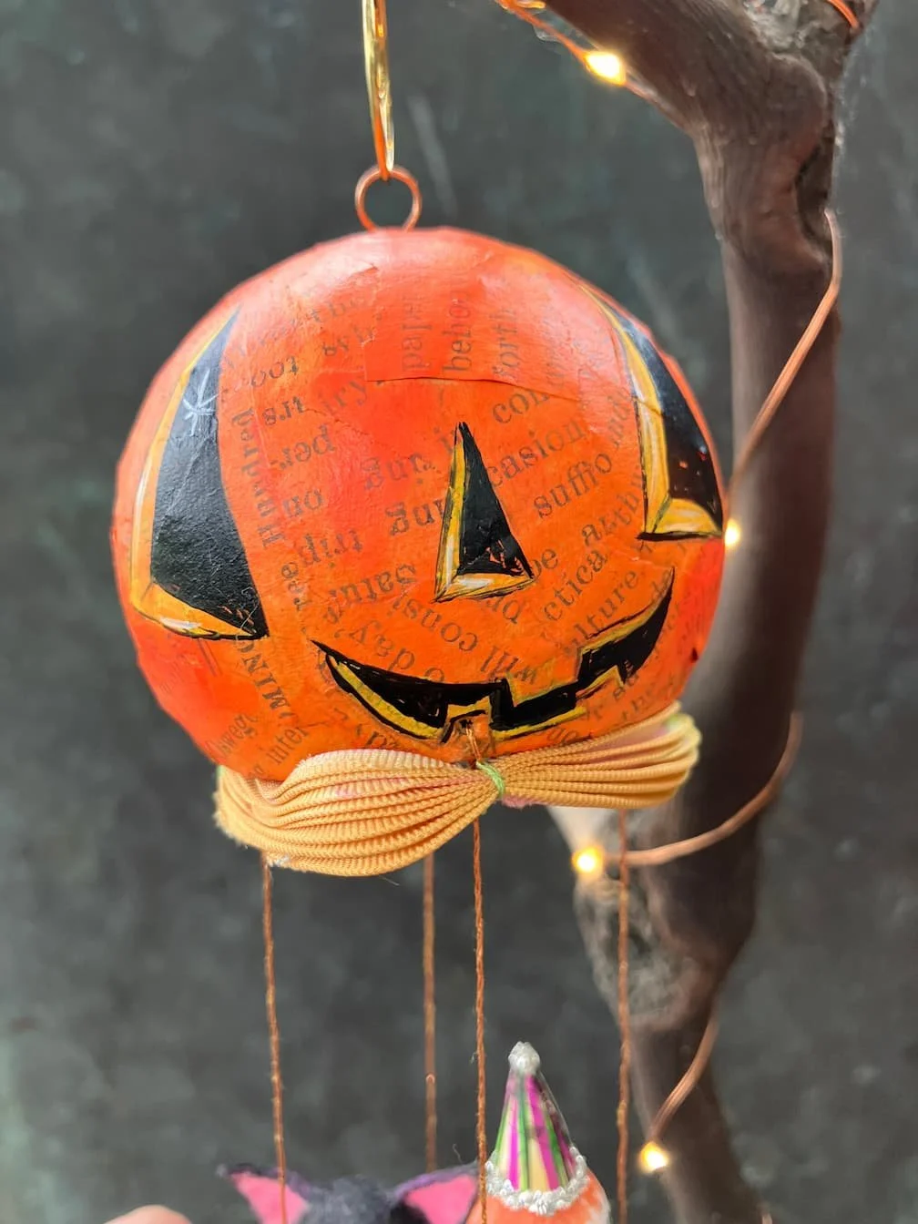 A Halloween-themed hanging decoration featuring a pumpkin with a carved face, painted with a black and orange design, and decorated with collage-style text and illustrations. It is suspended from a brown branch with string lights in the background.