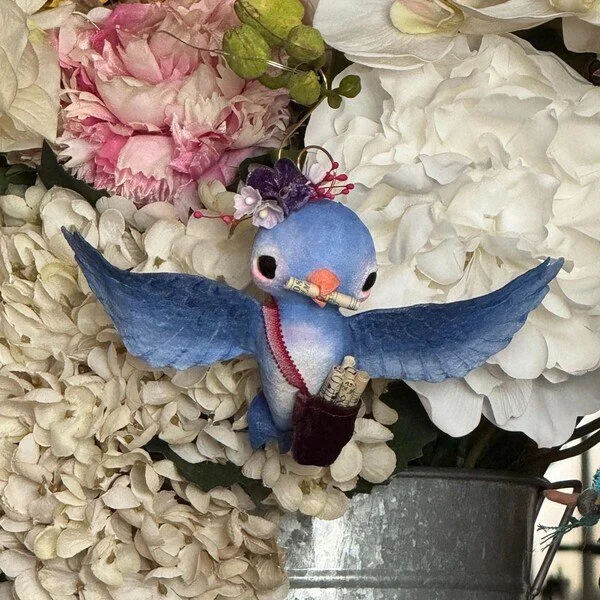 A decorative hummingbird figurine with blue wings, a purple crest, and an orange beak, holding a tiny book, surrounded by white and pink flowers.