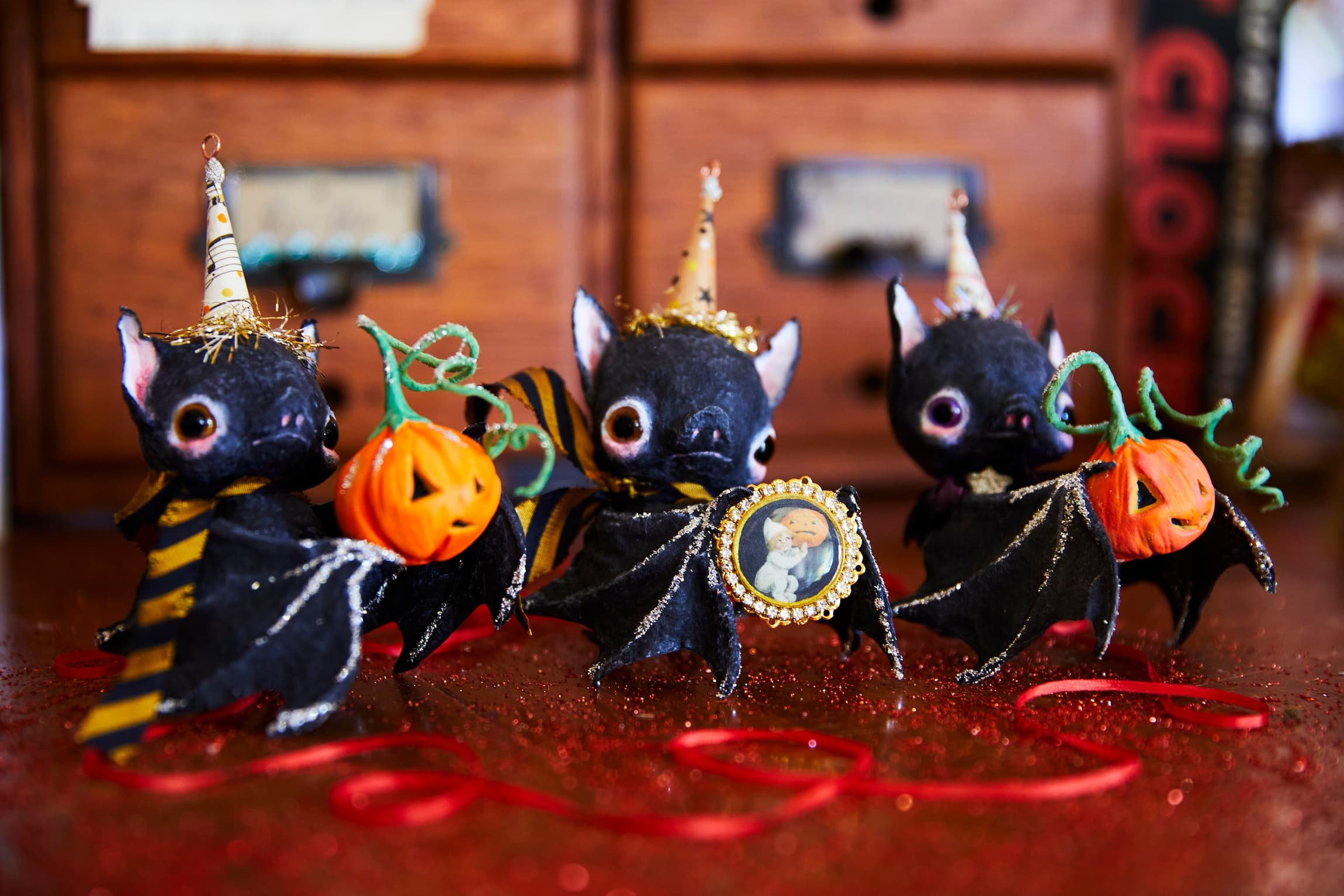Three spun cotton bat figurines dressed in black and gold costumes with Halloween-themed accessories, including pumpkin-shaped bags and striped wings, arranged on a sparkling red surface with a wooden dresser in the background.