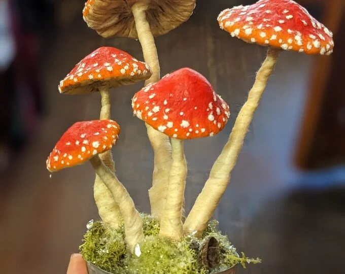 Artificial mushroom sculpture made from spun cotton with red caps and white spots, attached to beige stems, growing from a small pot with moss.