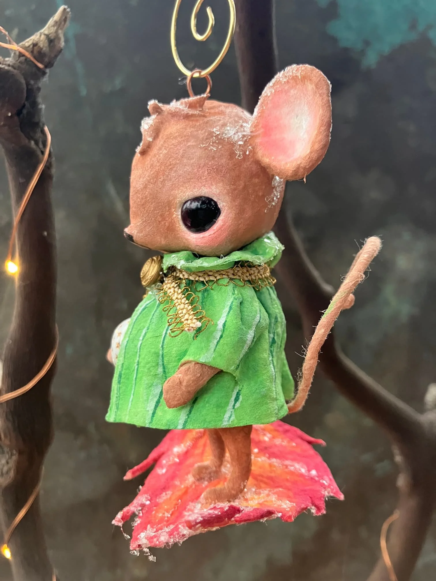 A small decorative mouse figure made of natural materials, wearing a green dress with a gold chain, standing on a red leaf-shaped base with pink accents.