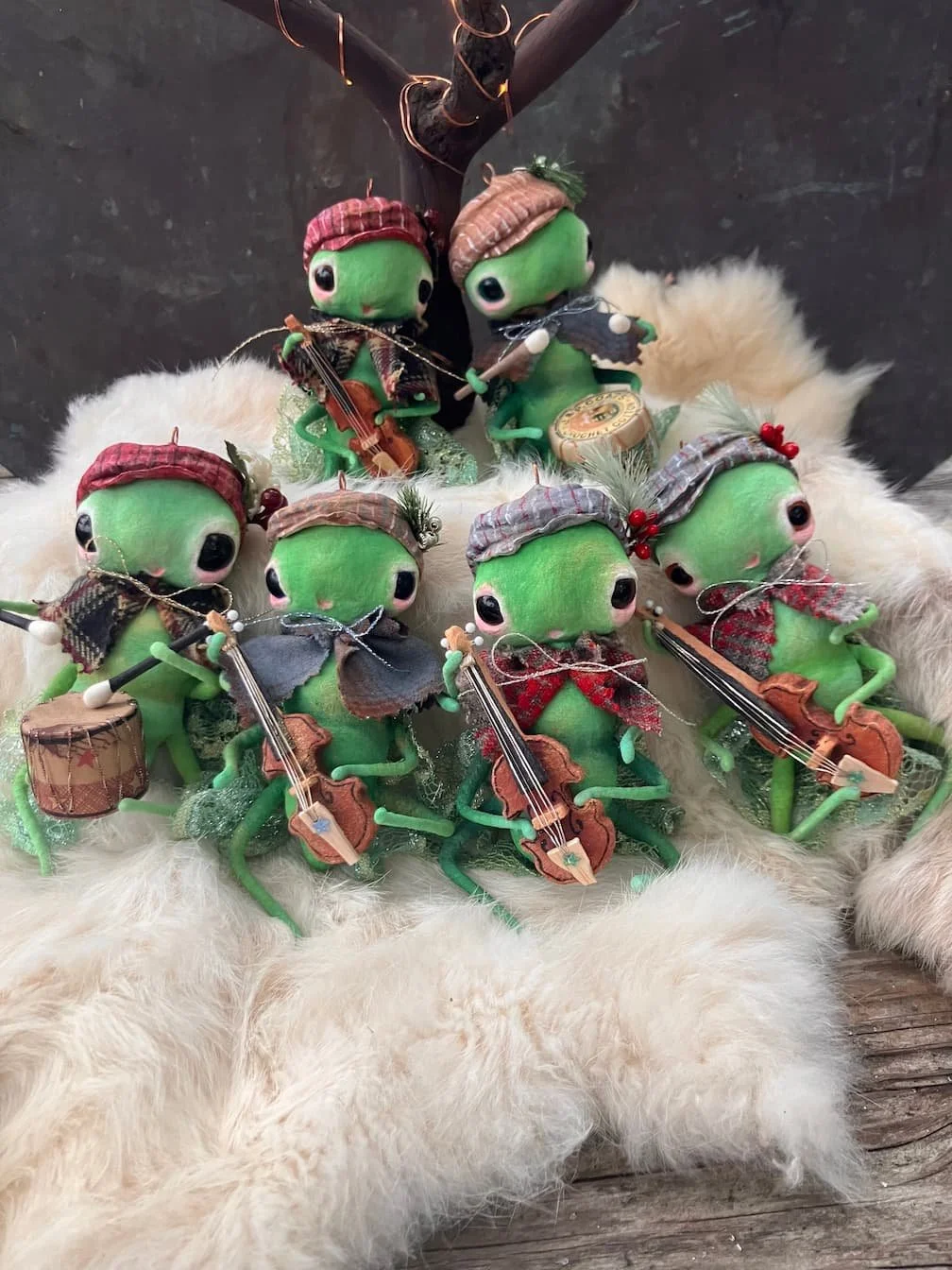 Set of six small green frog figurines dressed in plaid hats and scarves, playing tiny musical instruments including violins, drums, and a tambourine, arranged on a fluffy white surface with a dark background and a small faux tree.