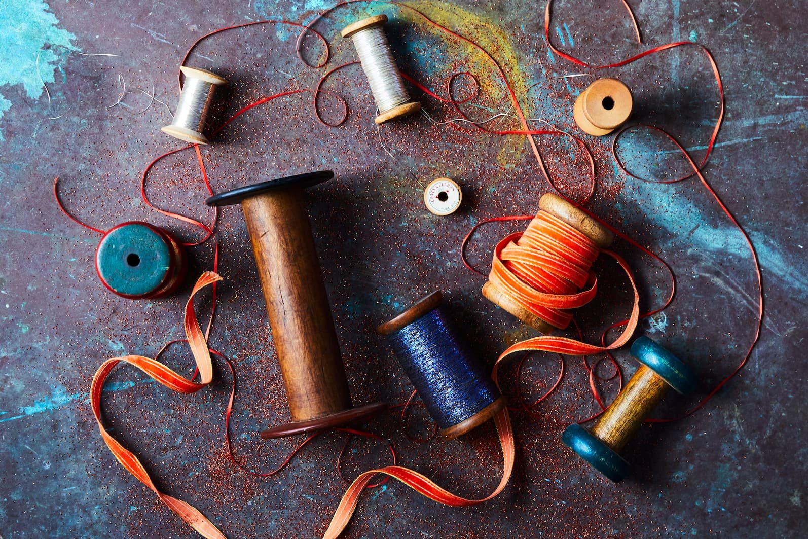 Colorful spools of thread and ribbons scattered on a textured dark surface, with some thread loosely wound around the spools and ribbons intertwined among them.