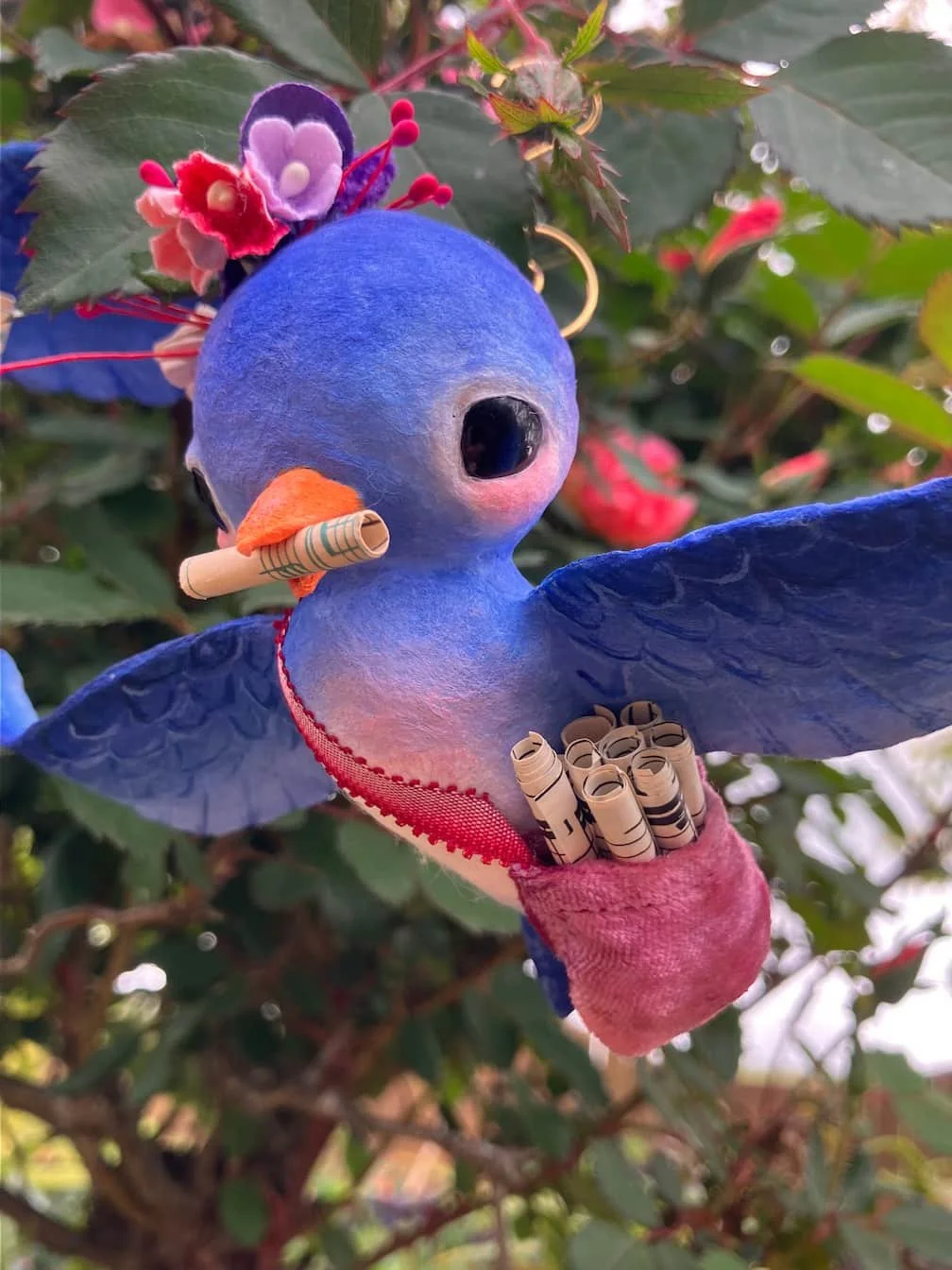 A colorful hummingbird-shaped ornament perched on a plant, adorned with flowers and rolled-up paper scrolls in its pouch.