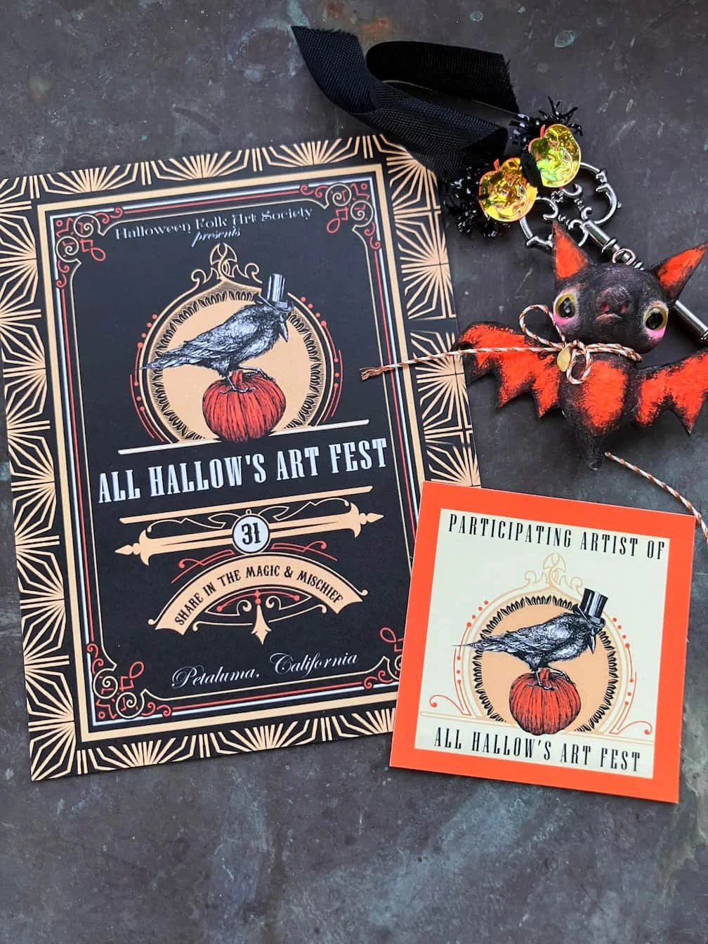 A Halloween-themed flyer for All Hallow's Art Fest, along with a small orange-bordered card, a keychain with a bat and a black cat with orange accents, and a decorative small bat figure with a black ribbon and yellow ornaments on a dark surface.