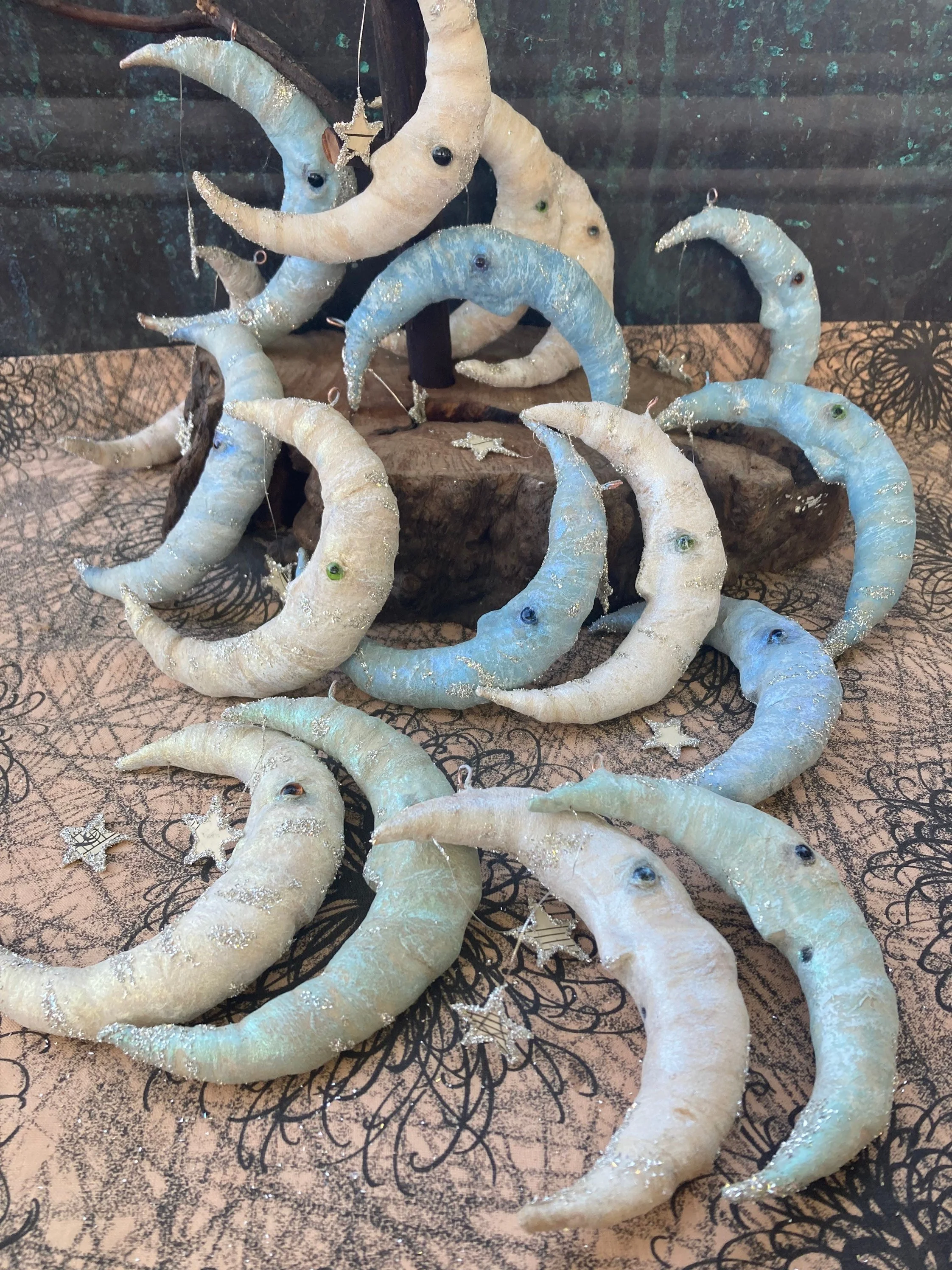 Multiple crescent moon-shaped ornaments with shiny accents and small googly eyes, displayed on a textured surface with a dark, leafy background.
