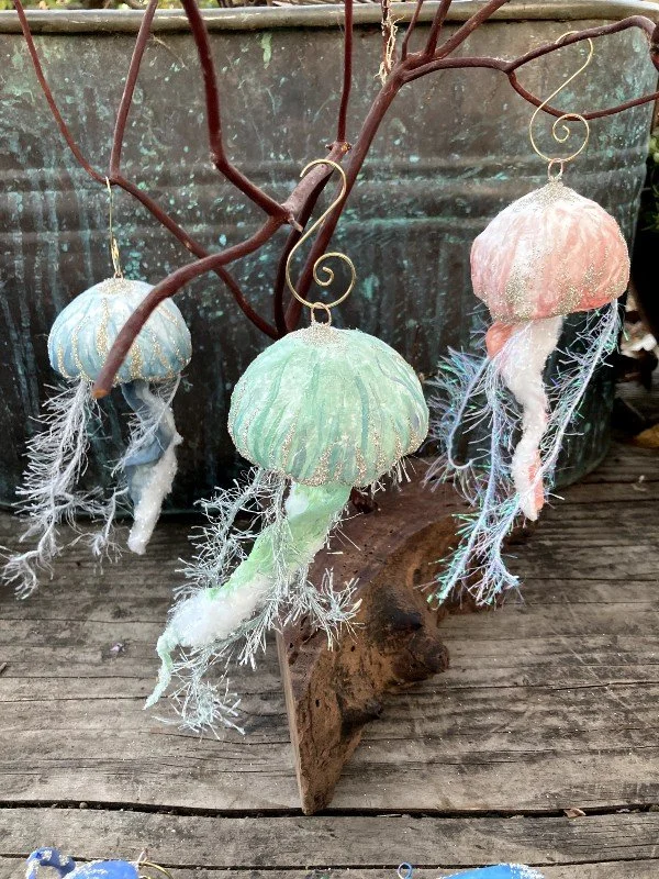 Three pastel-colored jellyfish ornaments hanging from leafless tree branches. The ornaments are made of spun cotton with fringed sparkling tentacles, and each has a metallic hook for hanging.