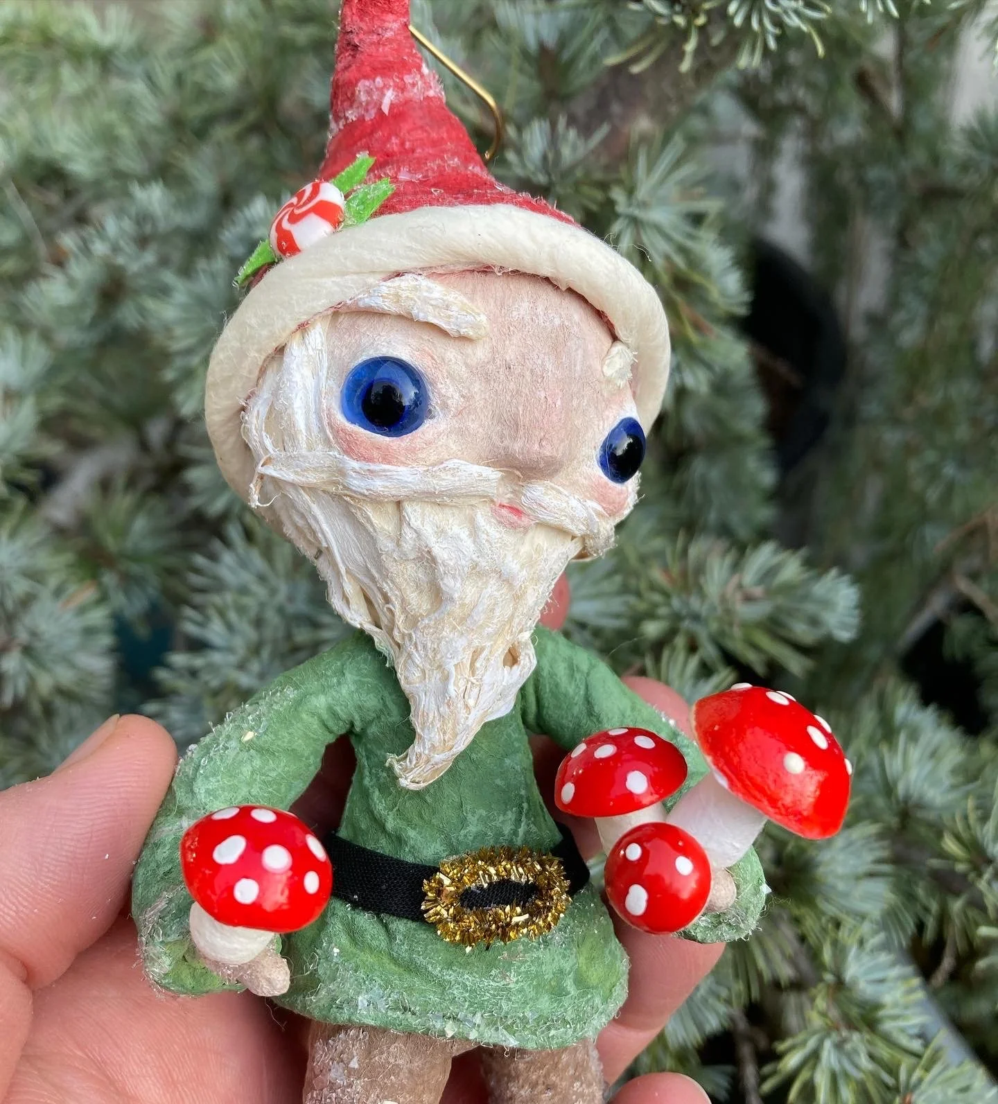 A Christmas tree ornament of a cartoonish elf with large blue eyes, white beard, wearing a red Santa hat and green outfit, surrounded by red and white spotted mushrooms.