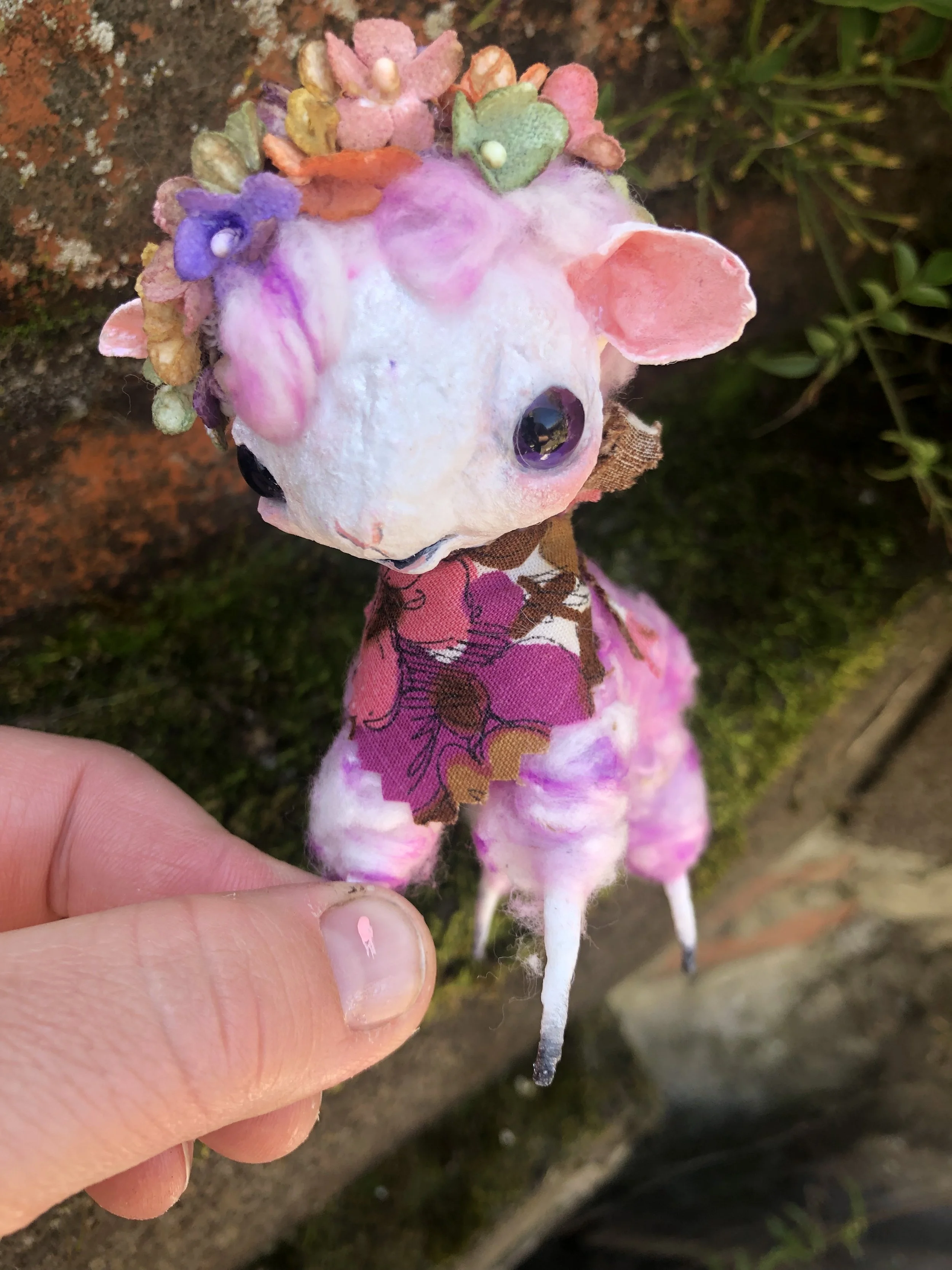 A small handmade ornament of a lamb with a white, fluffy body, pink ears, black eyes, and a pink patterned dress. The figure's head is decorated with a colorful felt flower crown.