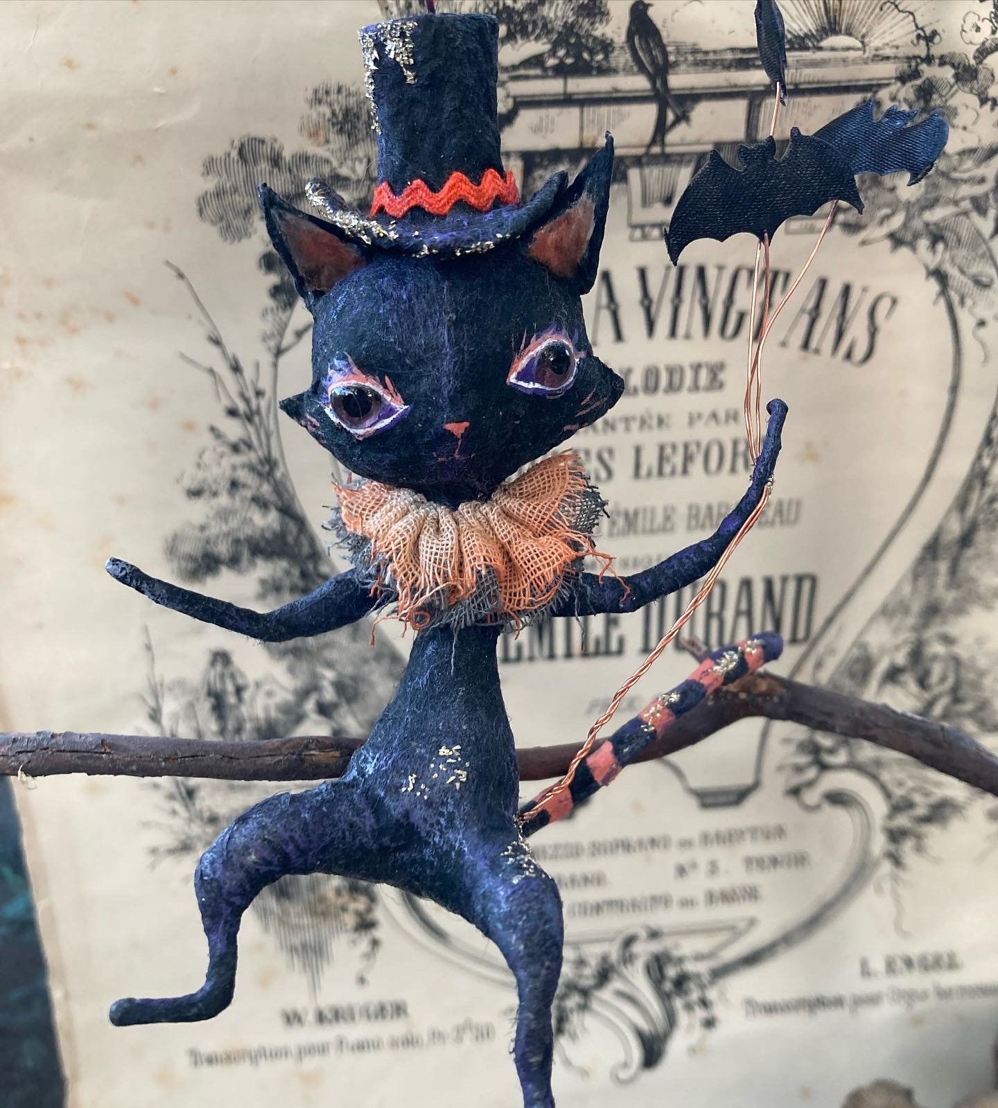 A whimsical figurine of a black cat wearing a top hat, orange ruffled collar, and holding a black bat, with a background featuring vintage-style text and decorative illustrations.