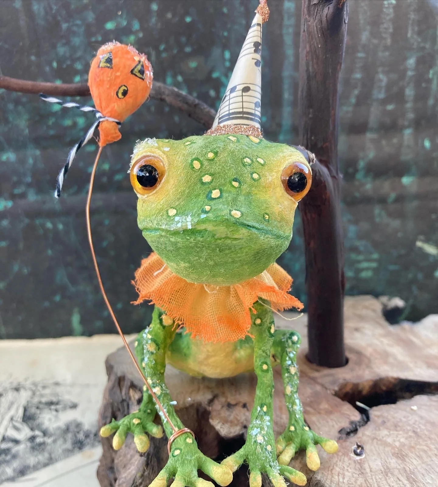 A handmade decorative frog with a green painted body and black eyes, wearing an orange collar and a cone-shaped party hat with musical notes. There is a small orange face with black features on a stick next to it, resembling a balloon or puppet, set 