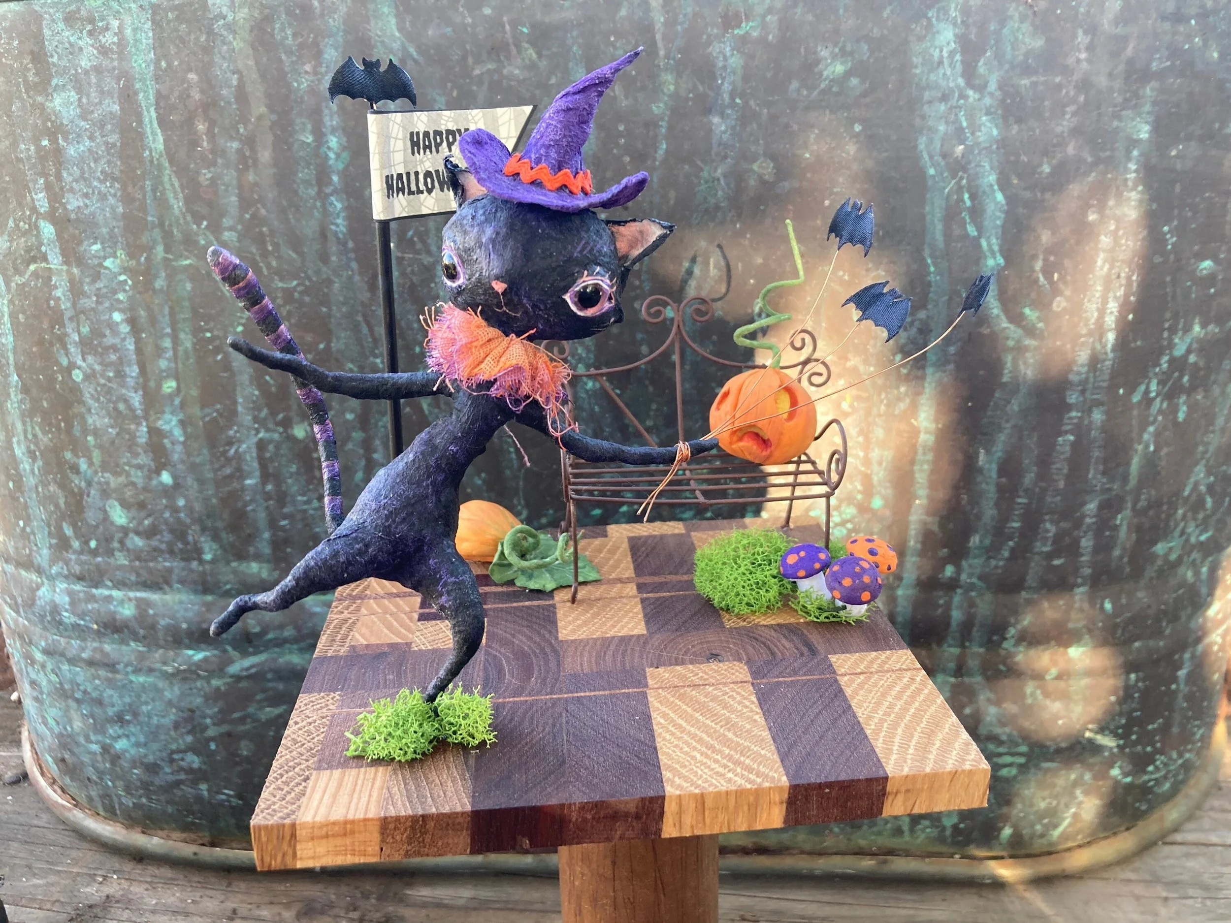 A Halloween-themed craft display featuring a black cat figurine with purple and orange decorations, a miniature pumpkin, toy bats, and artificial mushrooms on a wooden surface with a forest background.