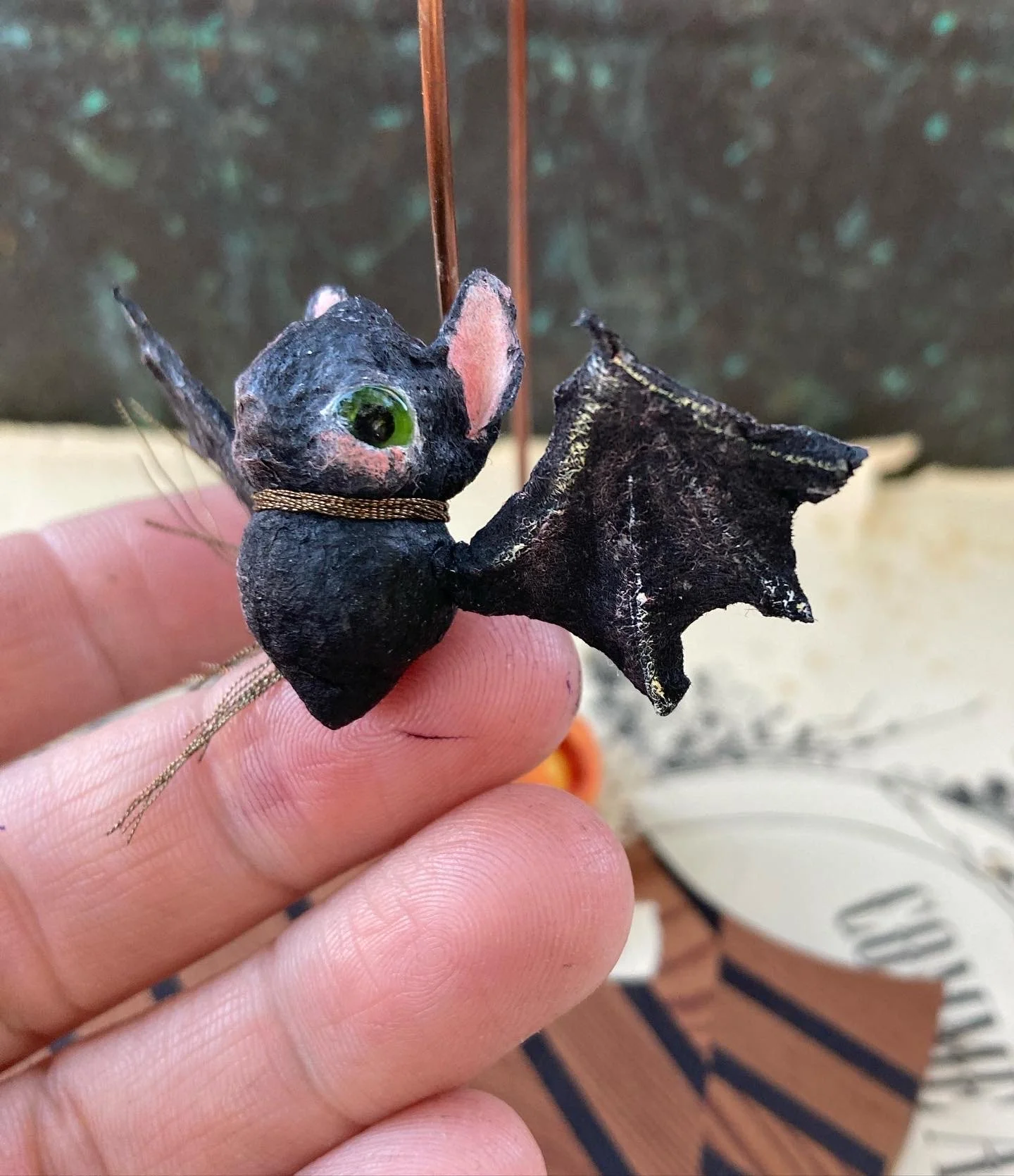 A small handcrafted bat figurine with a black body, green eyes, pink inner ears, and black bat wings. It is held by fingers and has a thin gold thread around its neck.