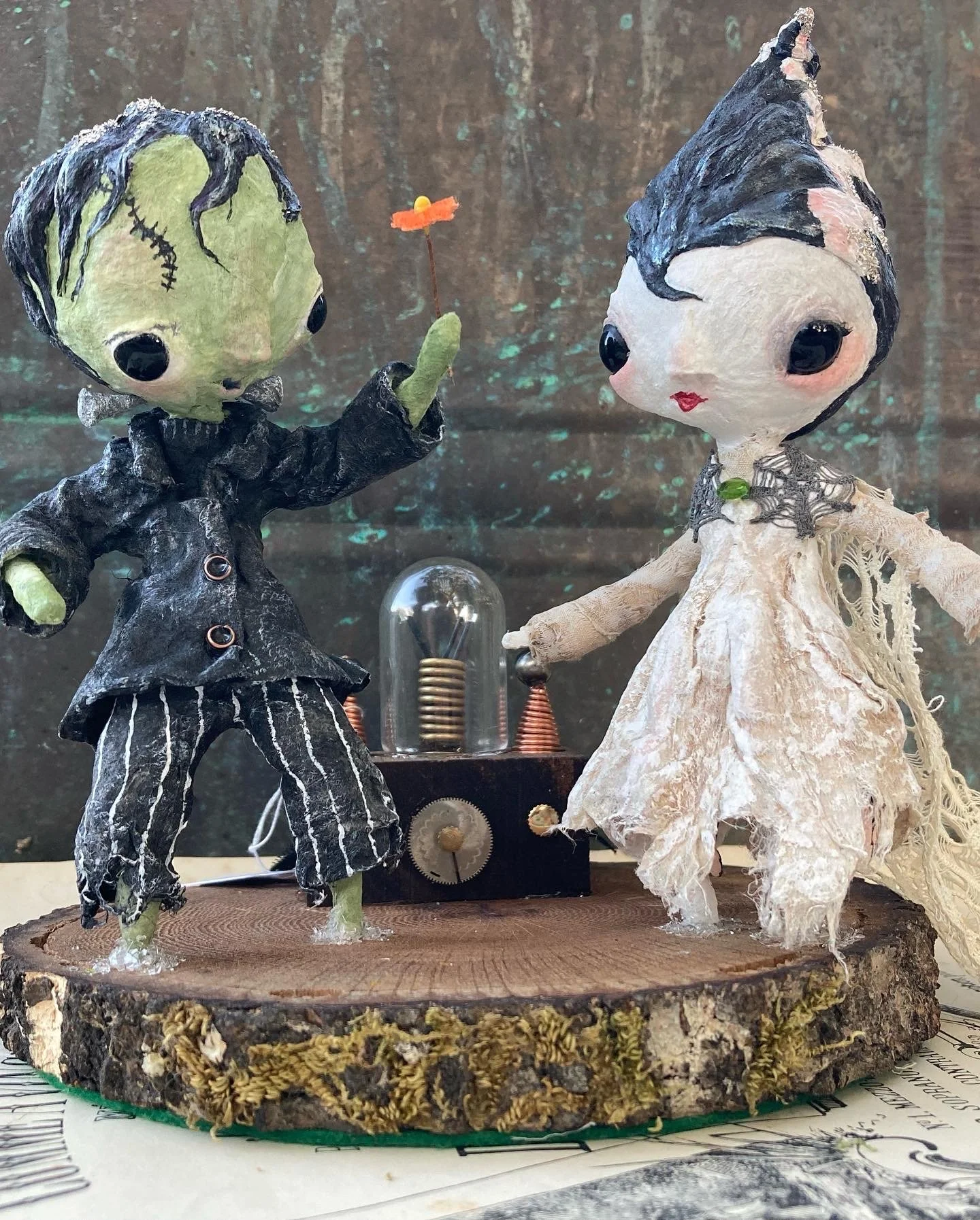 Two handcrafted dolls resembling zombies and a bride, standing on a wooden base inside a glass display case. The zombie doll wears a black jacket and striped pants, holding out a small orange flower with a yellow center toward the bride doll. The bri