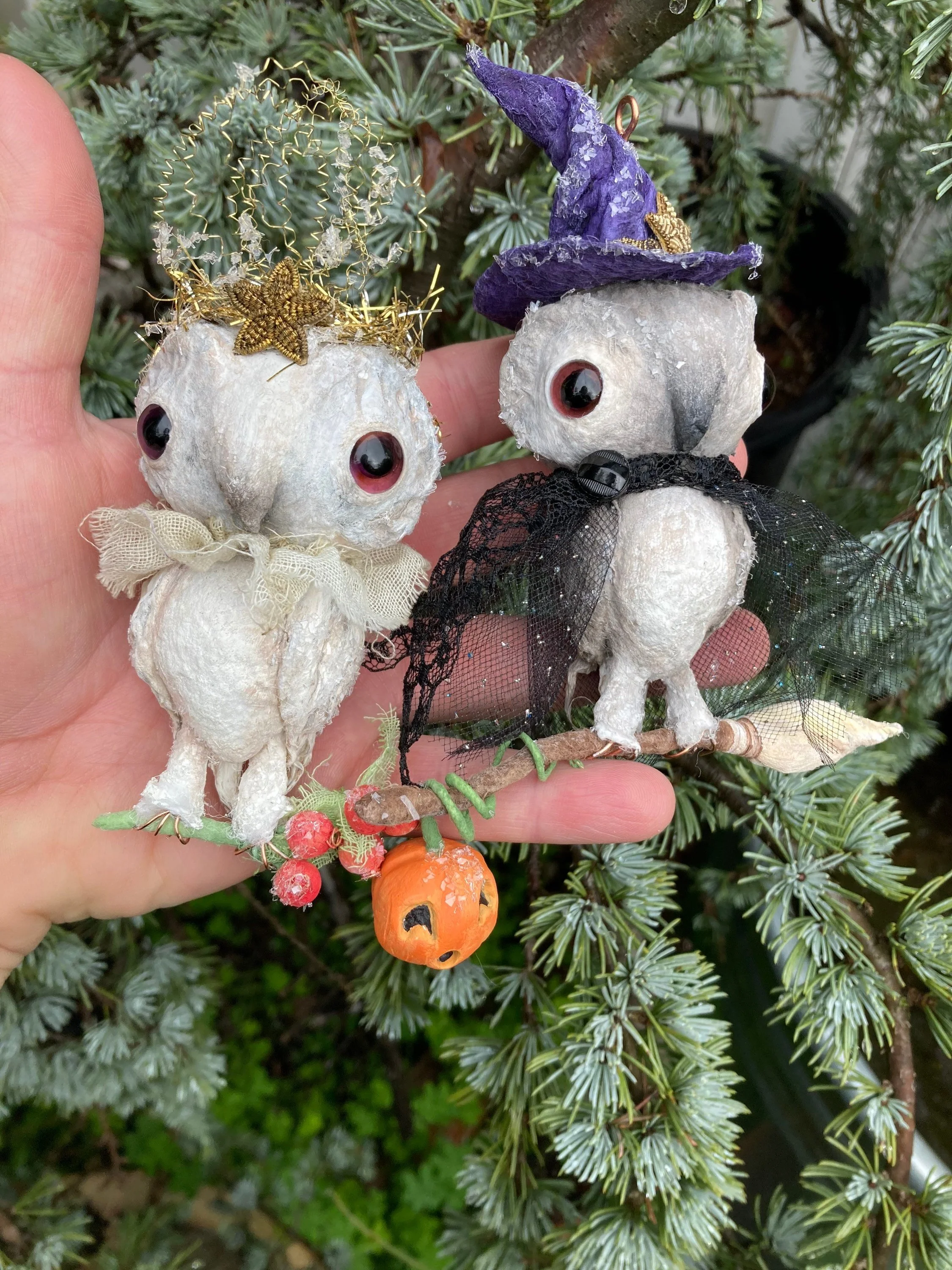 Two handmade Halloween decorations resembling skulls with large black eyes, one wearing a purple witch hat and the other wearing a black lace cape, held in front of a pine tree.