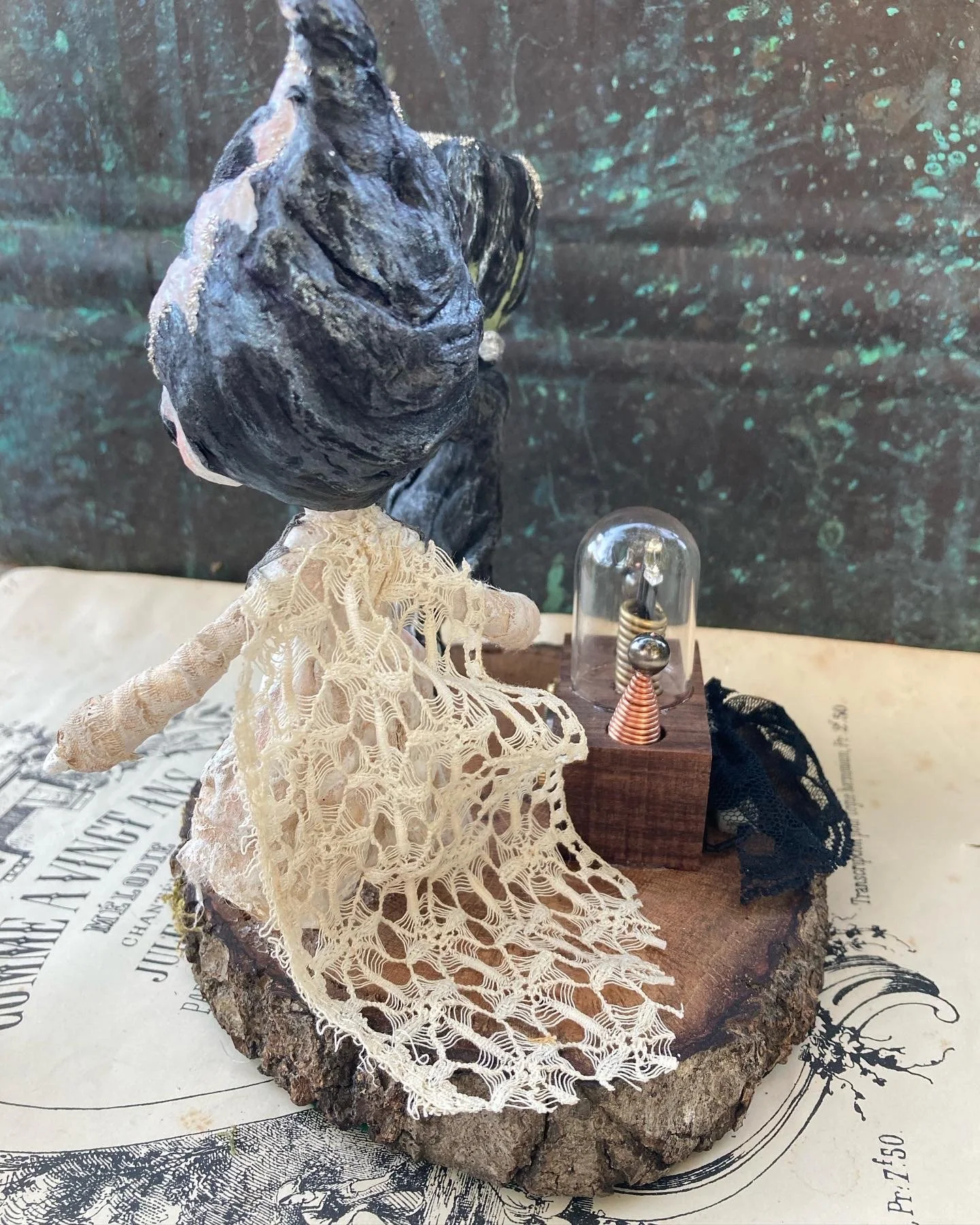 A mixed media art piece featuring a black volcanic rock with a white coral attached to it, a vintage light bulb encased in a glass dome, placed on a wooden block, with vintage lace fabric and black lace accents around it, set against a dark, sparkly 