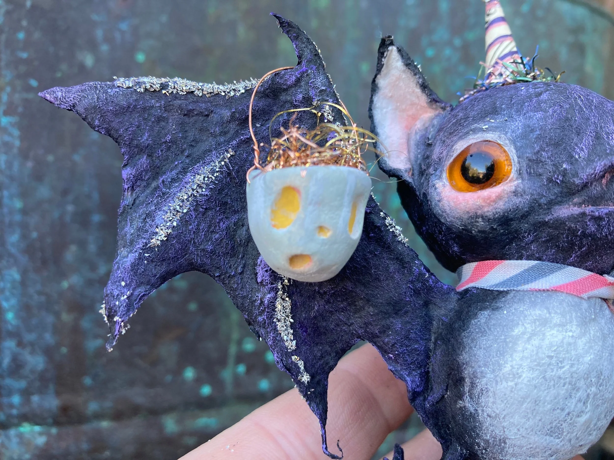 A person holding a bat ornament with purple accents, decorated with gold glitter, shaped like a cat's face with pointed ears and a face cutout, and a small pumpkin decoration with a lit candle inside attached to the hat.