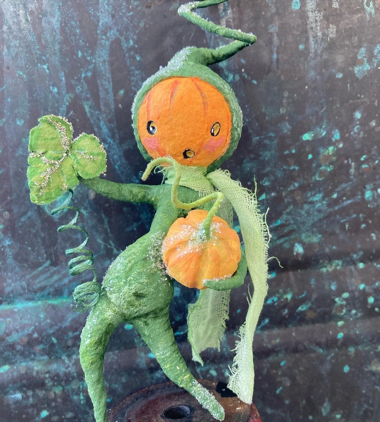 A mixed-media sculpture of a whimsical green creature with a face painted on an orange fruit, featuring a green vine tail, leaf-like arms, and fabric accents, set against a textured blue background.