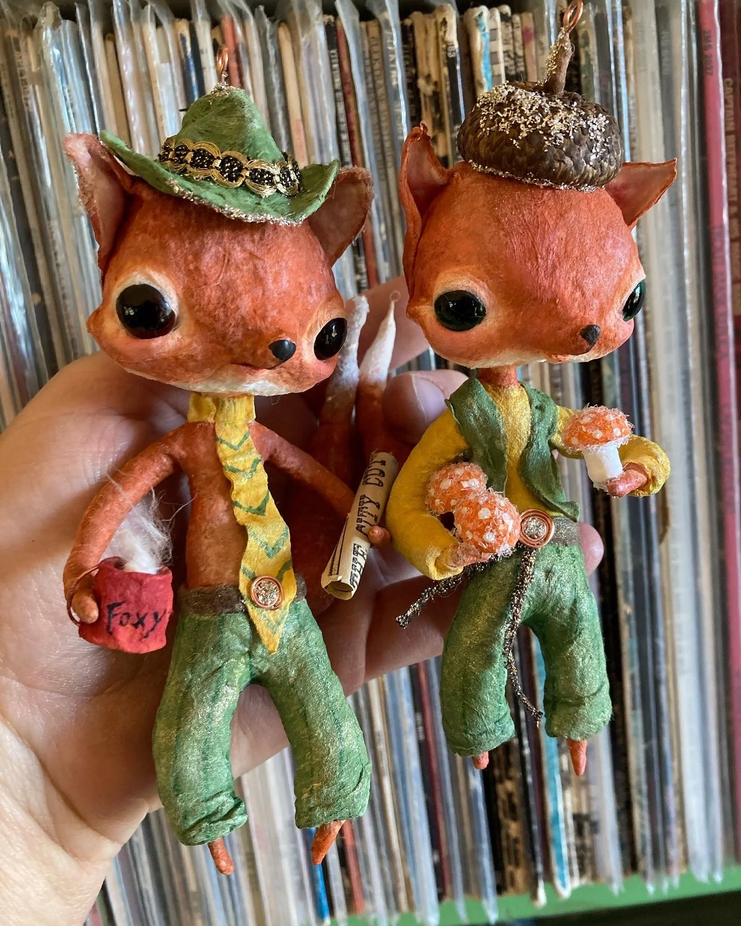 Two handmade fox figurines with large black eyes, orange heads, green bodies, and yellow and green clothing. Each fox wears a small hat: one green hat with black beads and trim, and a brown hat with white specks. One fox has a red cloth with the word