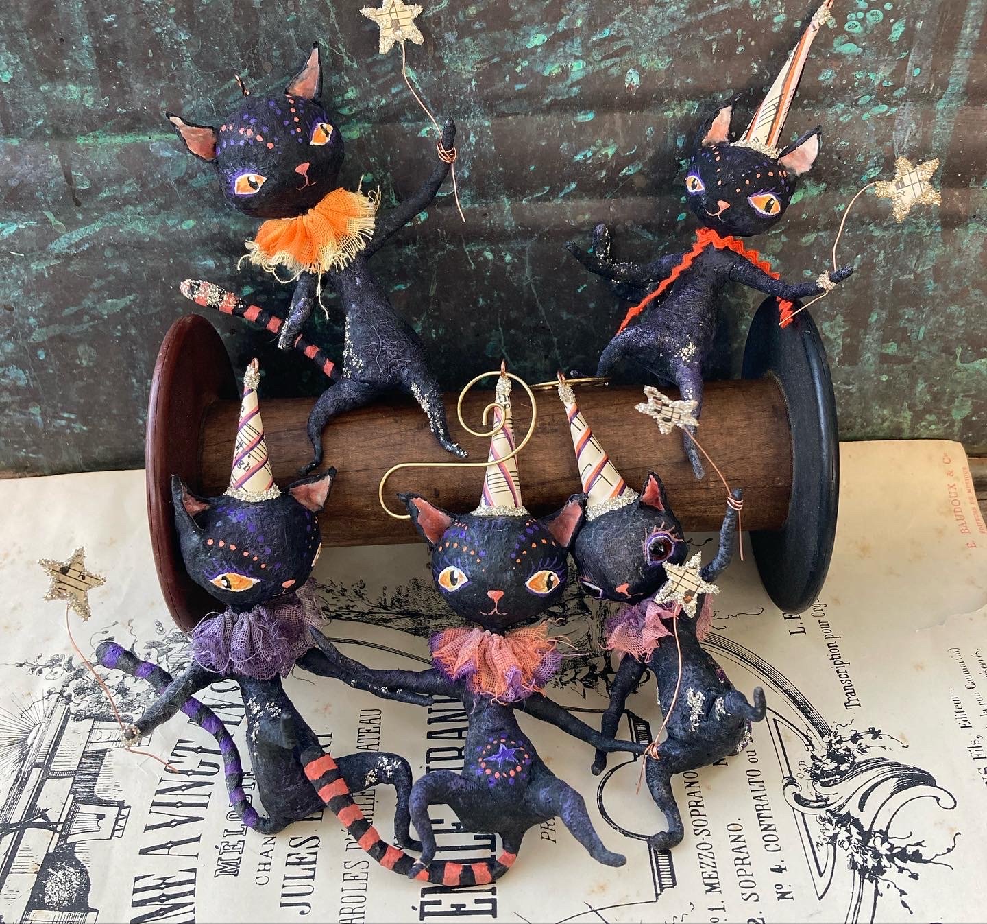 Multiple black cat figurines with various colorful details, wearing party hats and collars, arranged on a wooden spool, with some holding decorative wands and star-shaped sticks, placed on a sheet of vintage paper.