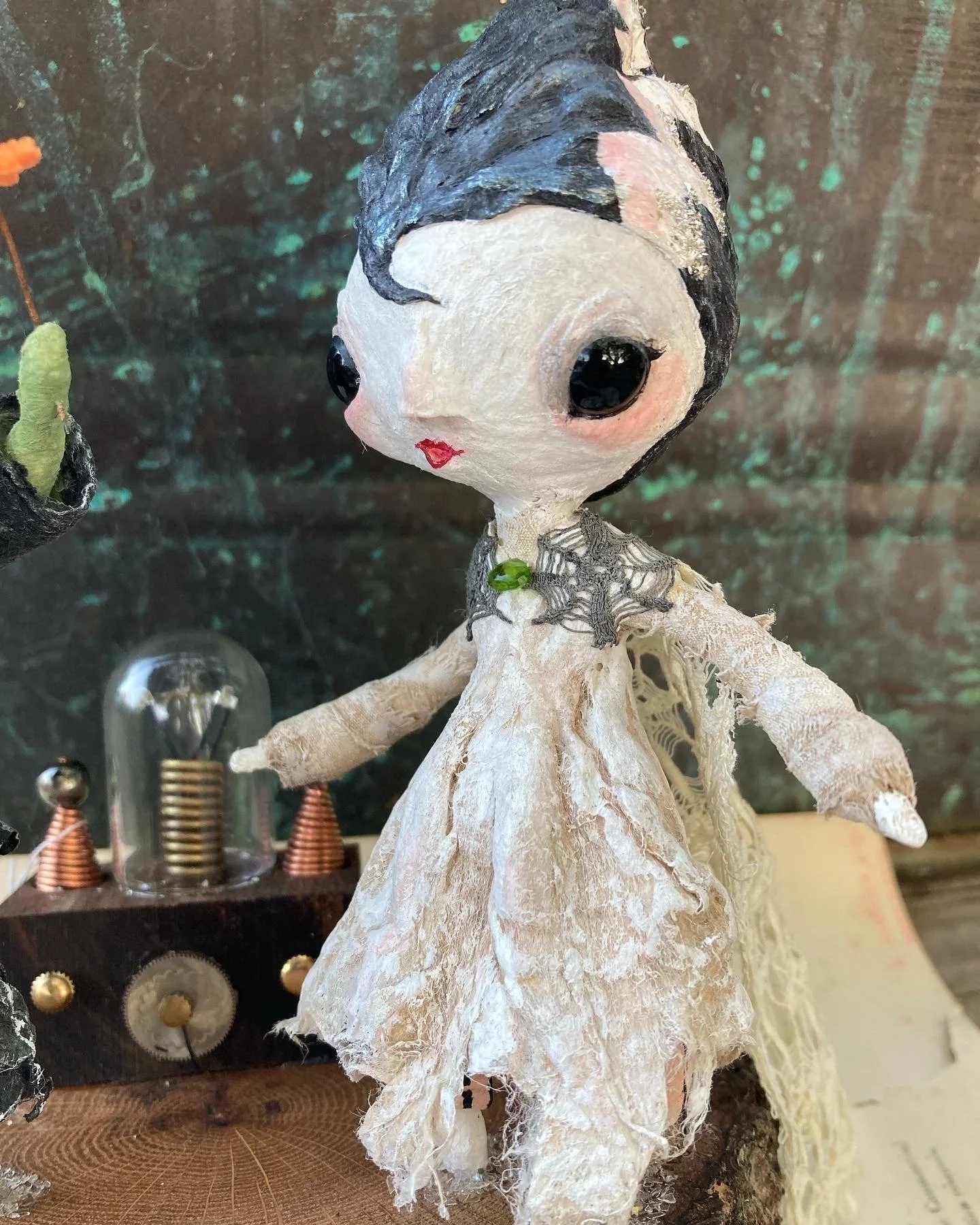 A doll-like figure with large black eyes, pink cheeks, and red lips, dressed in a gown made of textured, cream-colored material, with black hair and a necklace with a green bead, standing on a wooden surface with some copper-colored coil springs and 