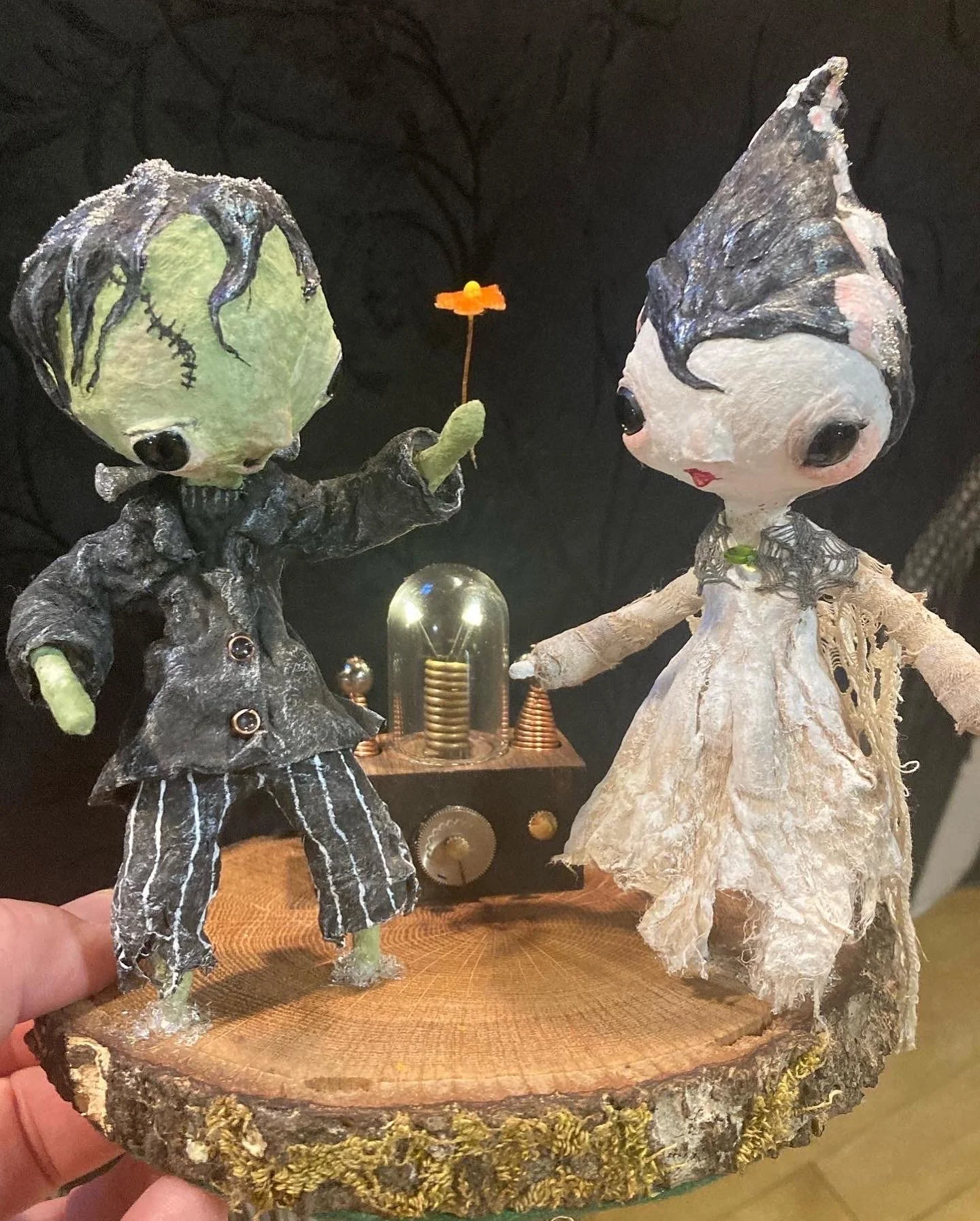 Two handcrafted dolls with large heads, dressed in vintage clothing, standing on a wooden base with a small scientific device in the background.