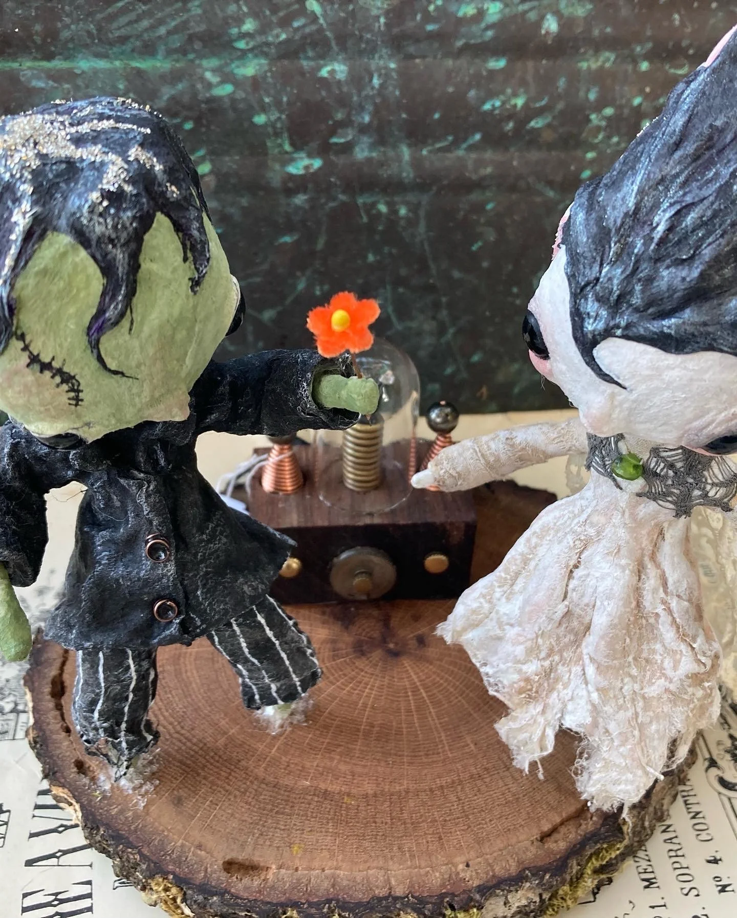 Two handmade dolls with dark and light color schemes are exchanging a small red flower with a yellow center. The dolls are placed on a wooden surface with a dark background.