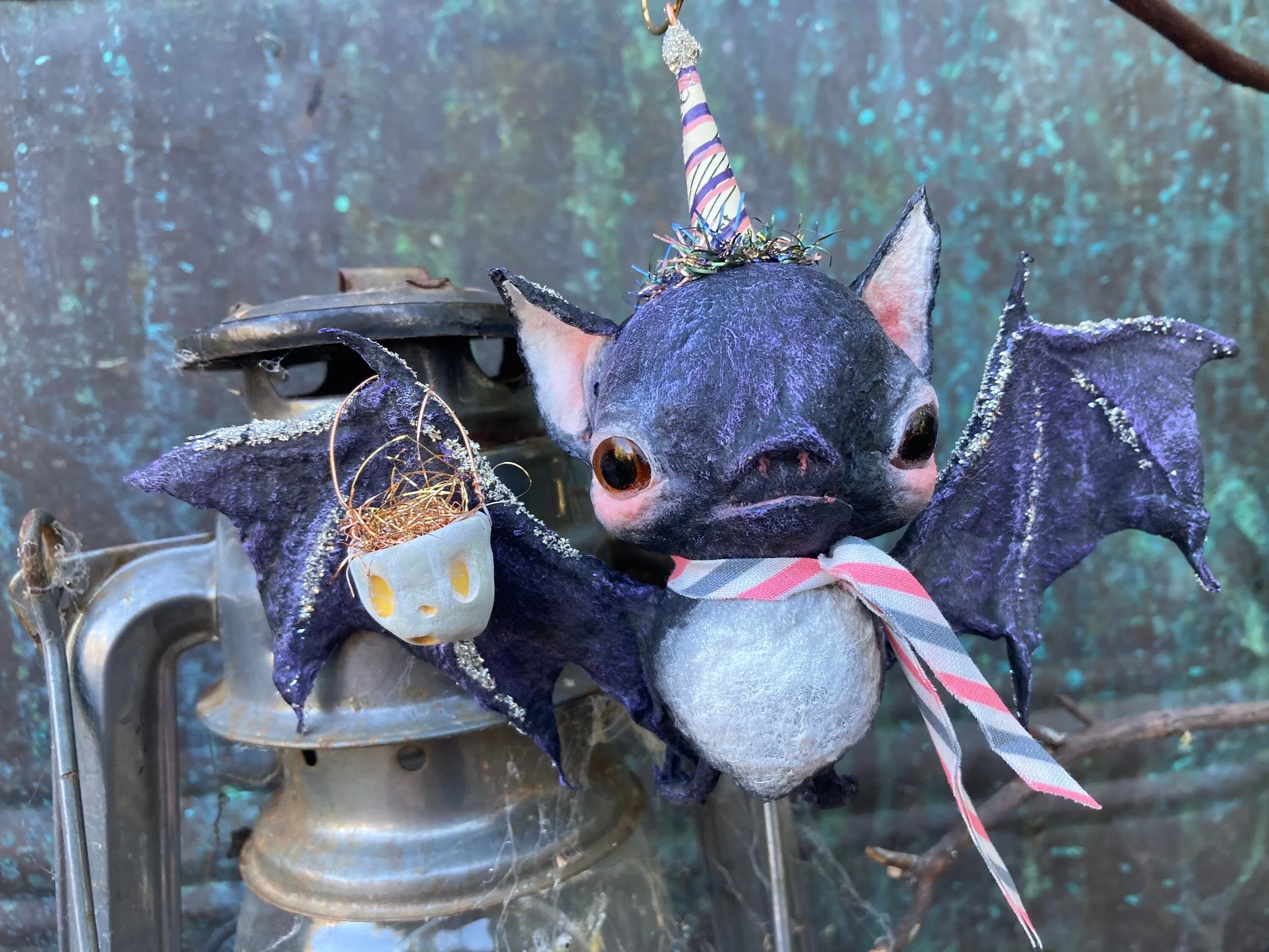 A sculpture of a cute bat with black wings and a snout, wearing a colorful party hat, with a collar of sprigs on its neck. The sculpture has large, hollow eyes and is mounted on a metal surface, with a textured blue-green background.