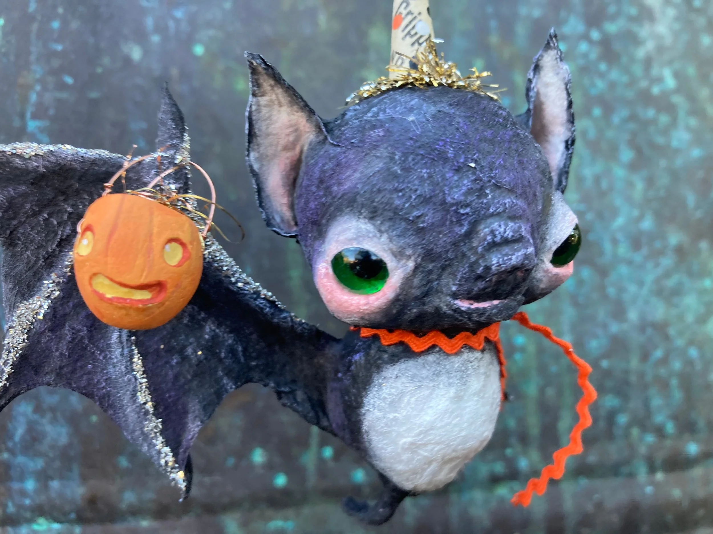 A Halloween spider decoration with a black body, green eyes, orange mouth, and orange mustache, wearing a white witch hat with gold tinsel, and decorated with glitter and red string, against a blurry blue background.