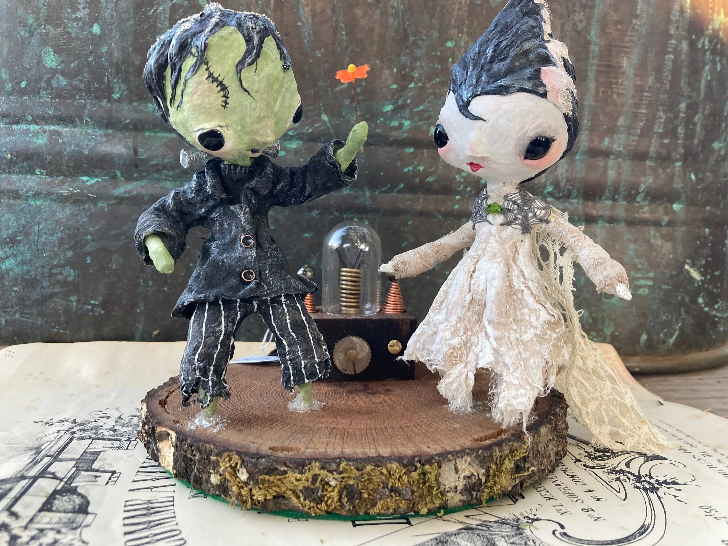 Two handmade figurines of a male and female are standing on a round wooden base. The male figure has green skin, black and purple hair, and is wearing a black jacket with white striped pants. He is holding a small orange flower with a yellow center. 