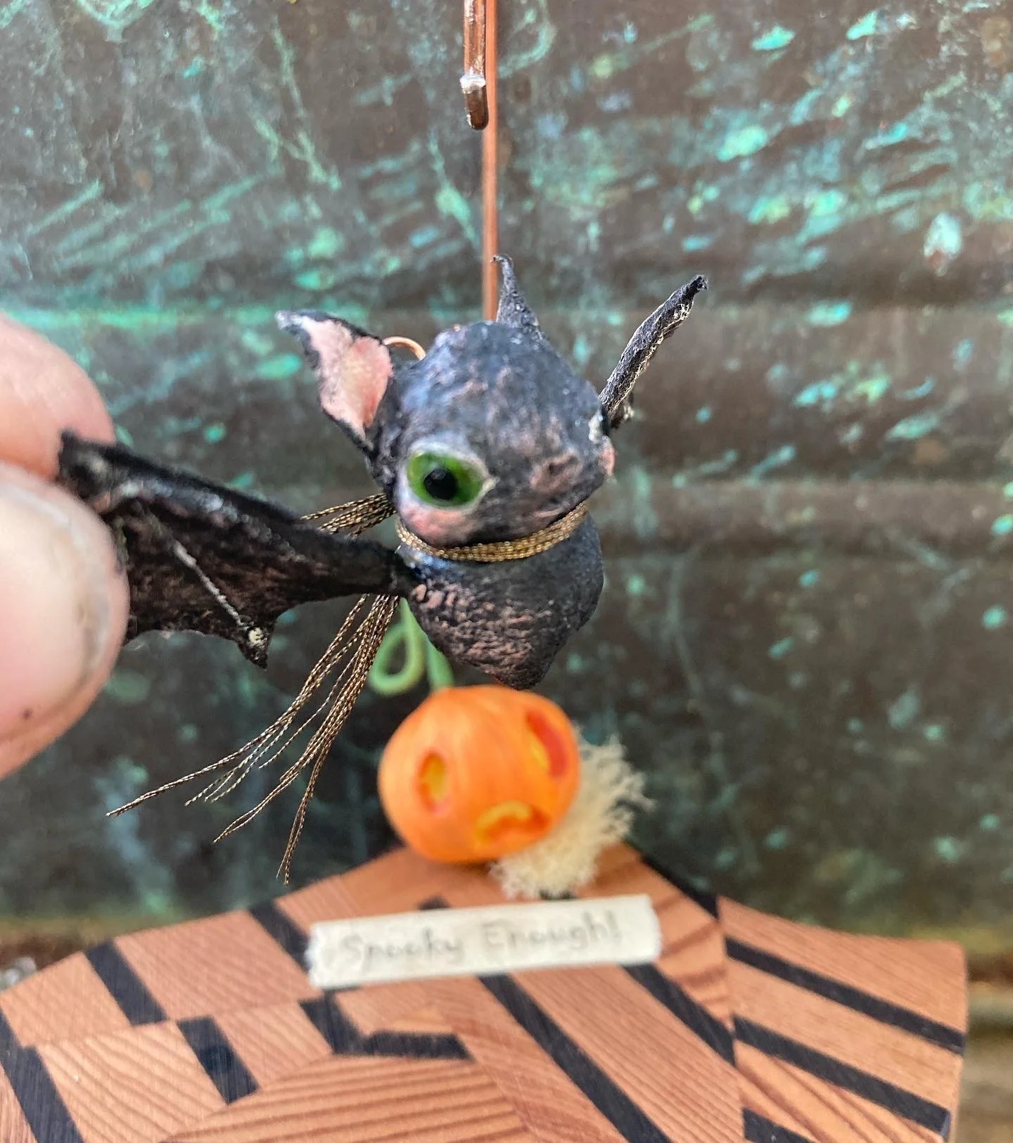 A small, handmade Halloween decoration featuring a black cat with large green eyes, fuzzy ears, and detailed wings, attached to a piece of wood with a sign that reads 'Stress Enough!'. In the background, there is a small orange pumpkin with a carved 