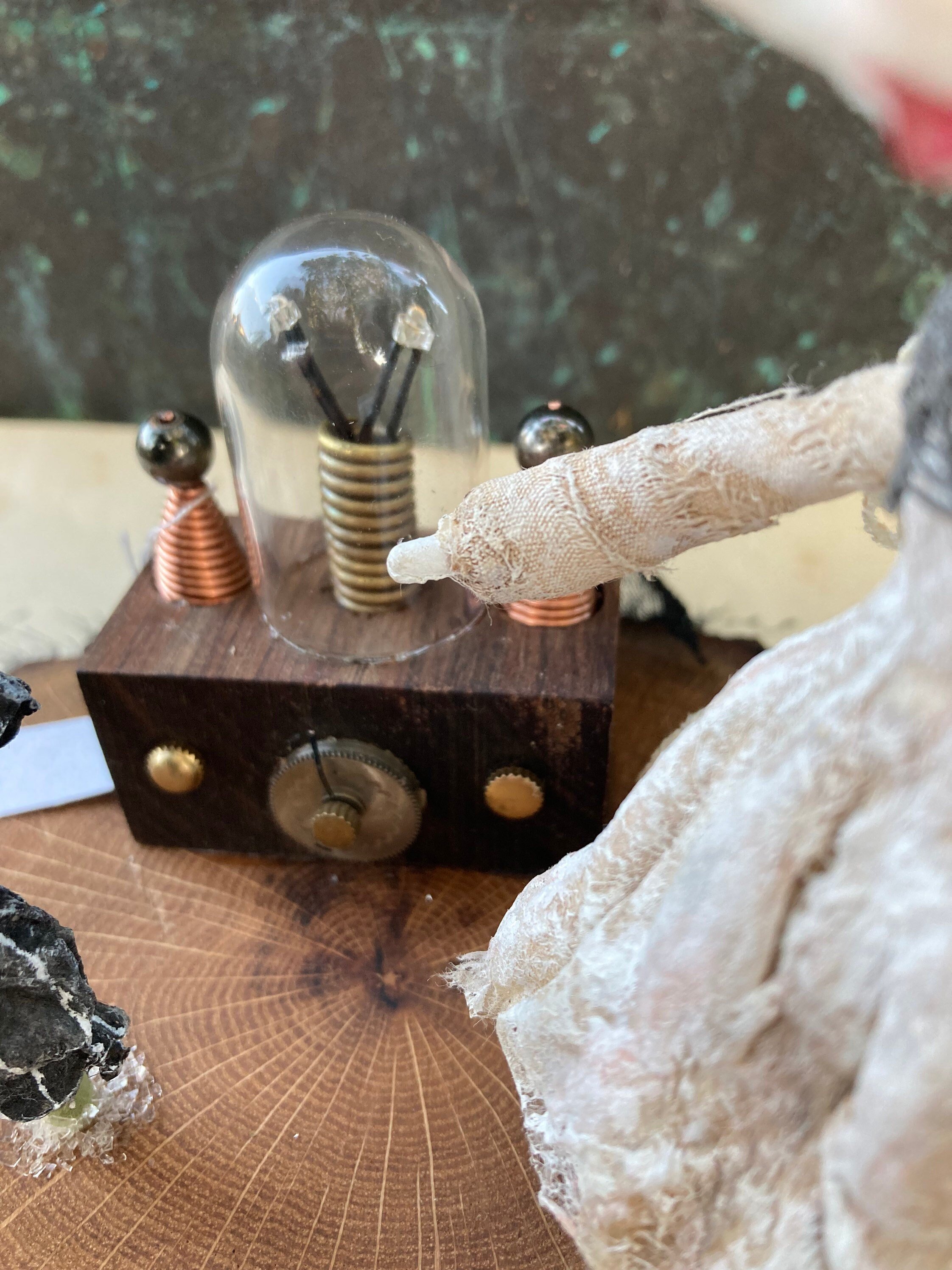 Close-up of a small sculpture with vintage-style electrical device with glass and copper components on a wood surface, surrounded by textured objects and a piece of crumpled tissue paper.