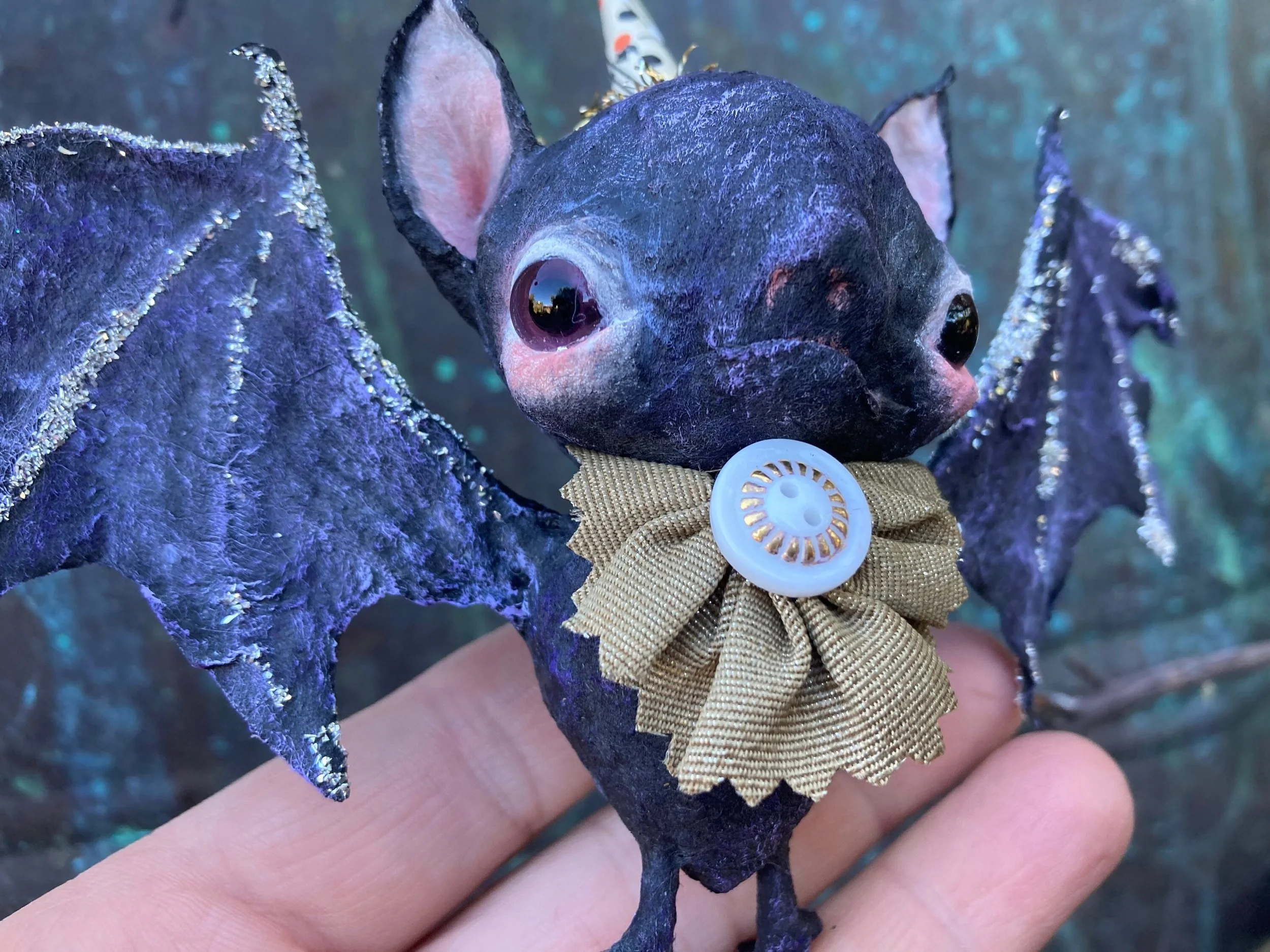 Close-up of a hand holding a cut bat ornament featuring pastel pink inner ears and a cream-colored bow with a decorative vintage button.